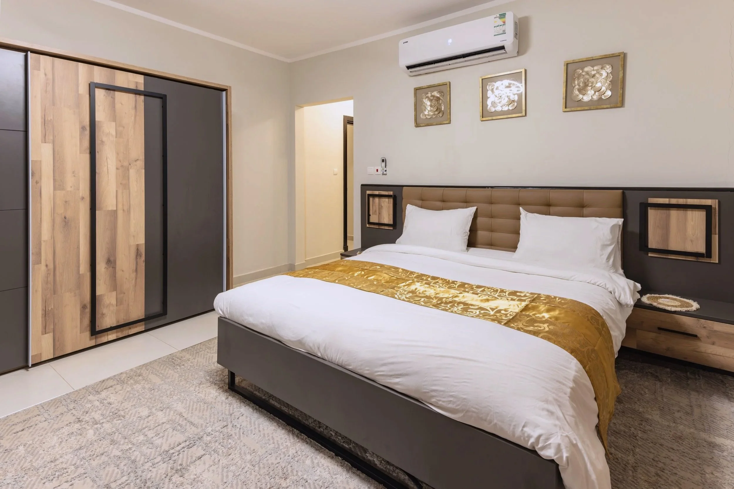 Modern bedroom with a large bed, gold decorative runner, beige headboard, wall-mounted air conditioner, artwork on the wall, and a sliding closet door.