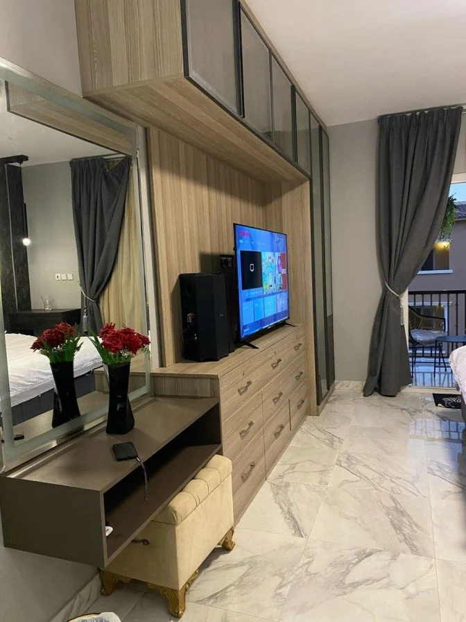 Living room with a wooden entertainment center, flat-screen TV, black speaker, and a remote. A mirror reflects a portion of the bedroom with a bed and a vase of red flowers. There are dark curtains and a balcony with outdoor furniture.