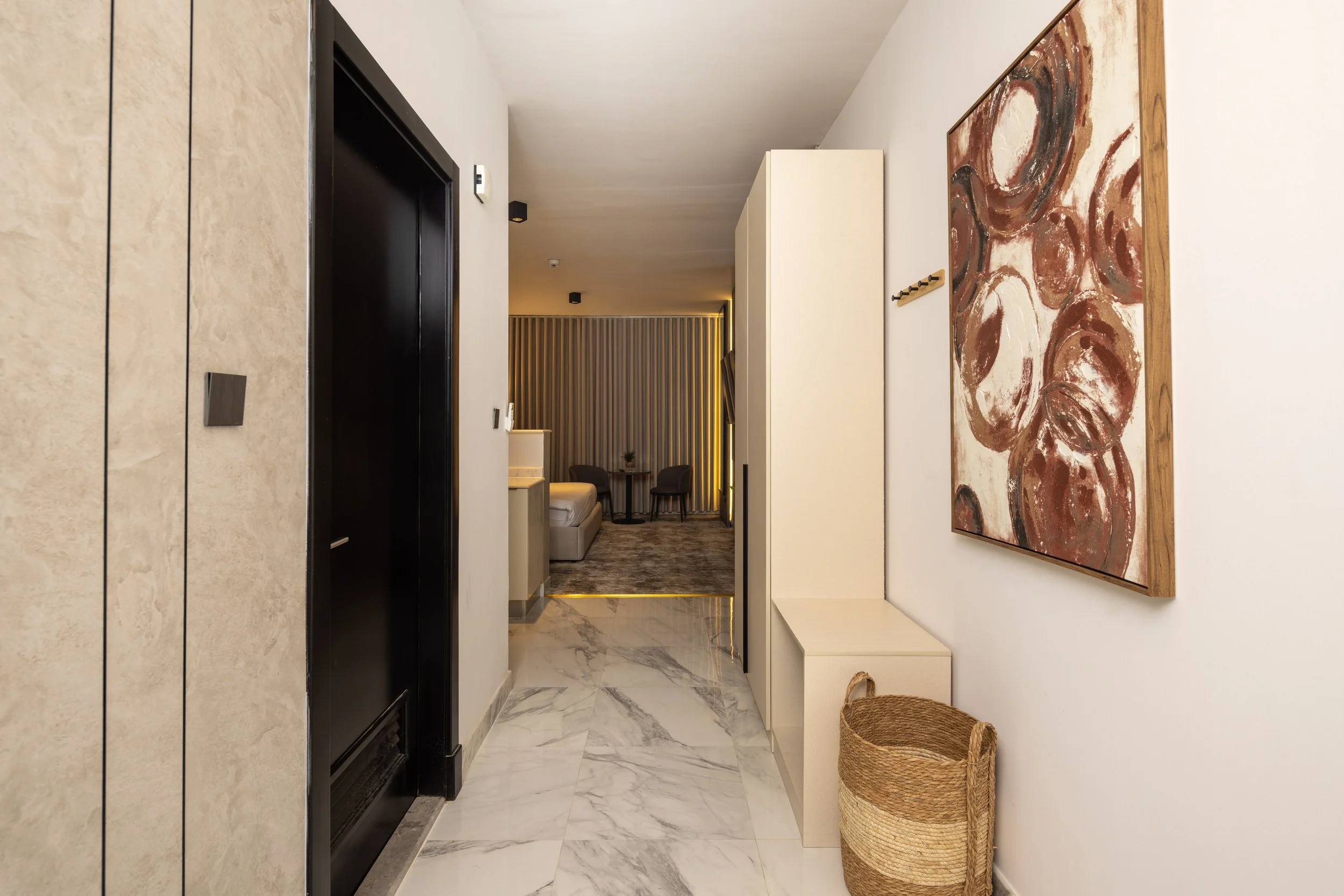 Living room with beige sofa, dark and patterned pillows, a large abstract painting, a tall potted plant, a brown leather chair, a glass table with flowers, and a sliding glass door leading to a balcony.
