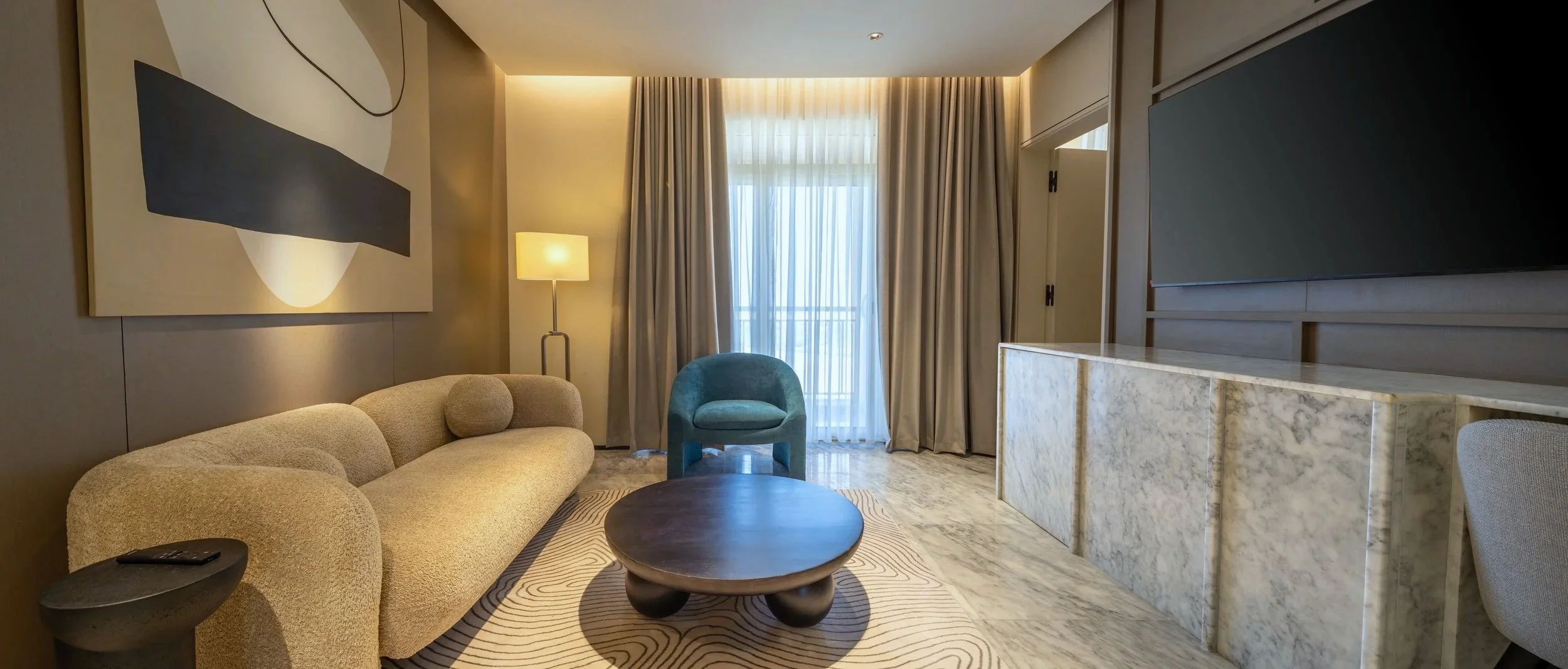 Modern hotel living room with beige sofa, blue armchair, round coffee table, marble sideboard, TV, floor lamp, large window with beige curtains, and abstract wall art.