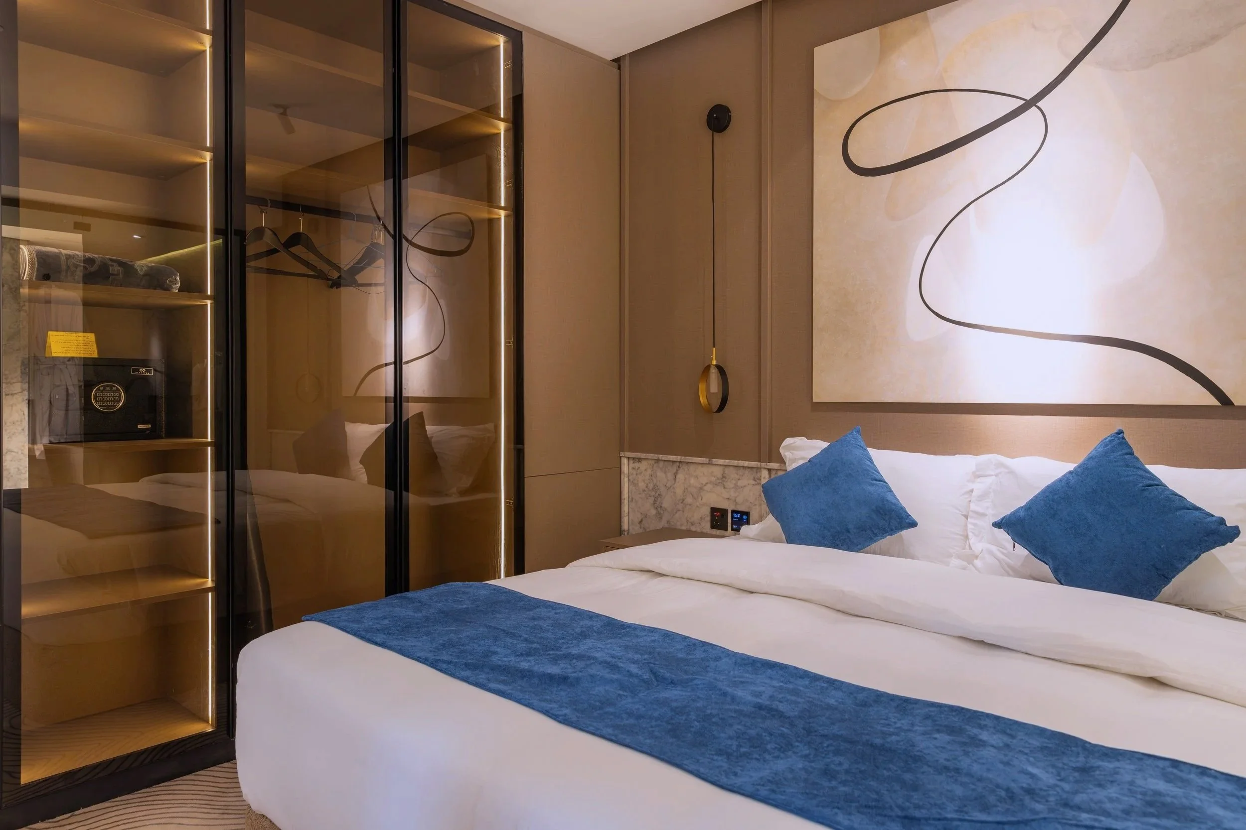 A modern hotel room with a large bed, blue accent pillows, and a matching blue bed runner. There is a large abstract wall art above the bed, and a pendant light hanging beside it. To the left, there is a built-in closet with glass doors showing shelves and a safe inside.