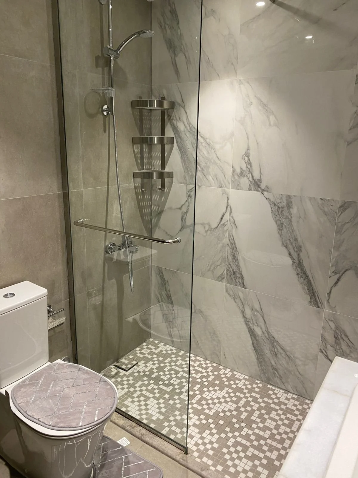 Modern bathroom with a glass-enclosed shower area, marble wall tiles, a handheld showerhead, three metal shelving baskets, a white toilet with a decorative towel, and a beige tiled floor.