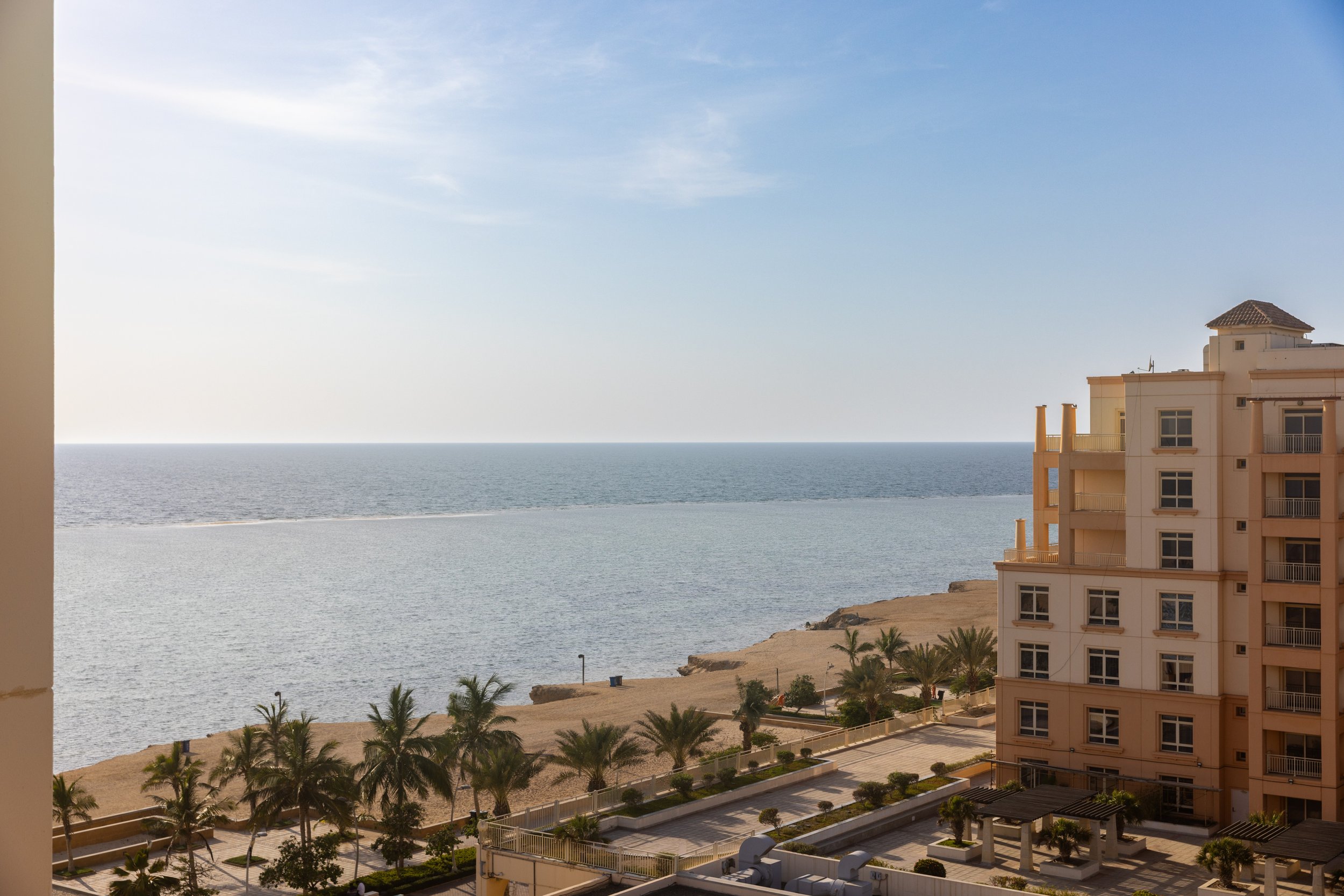 View of the ocean and sandy beach from a balcony, with a multi-story beige building, palm trees, and a partly cloudy sky.