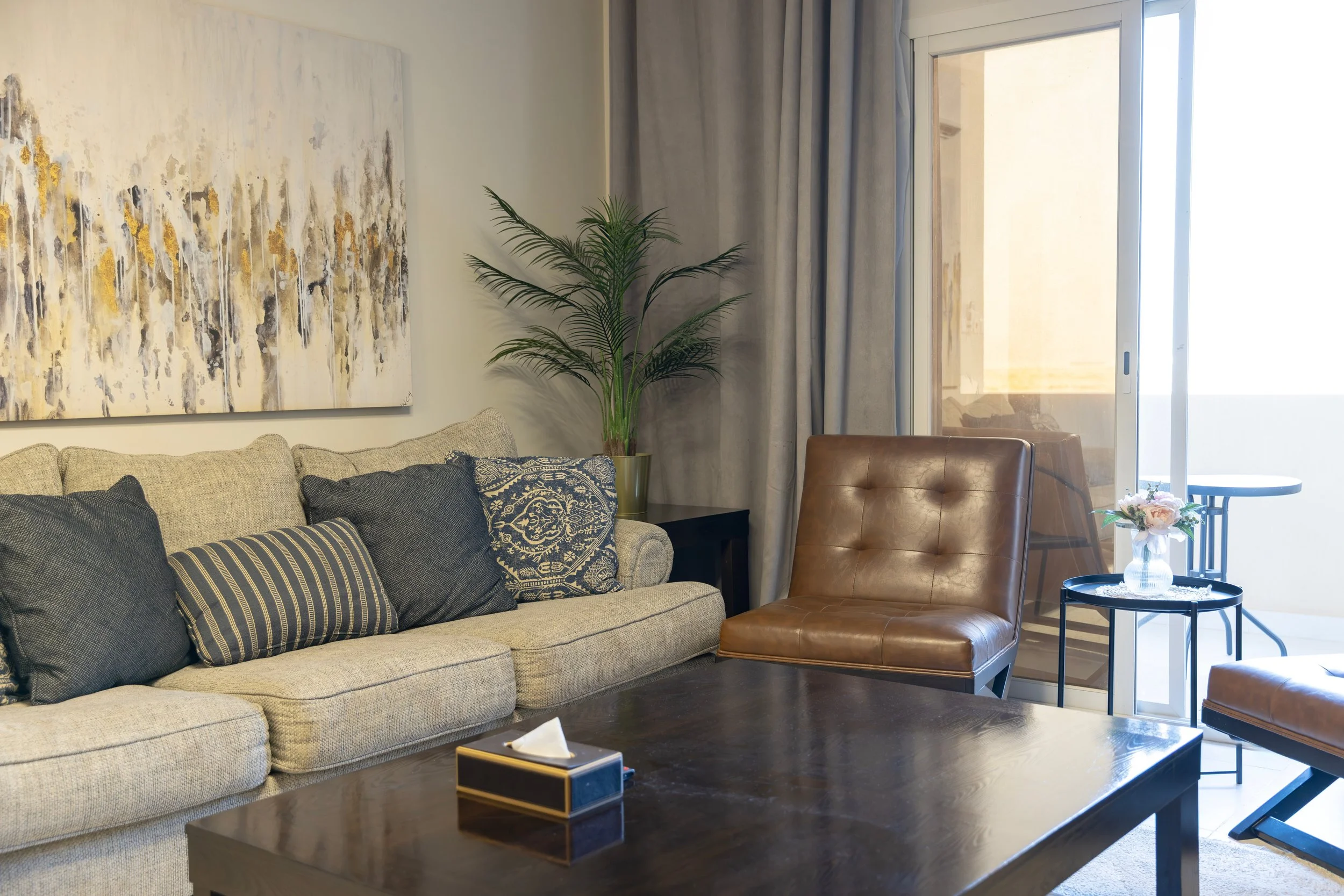 Living room with beige sofa, dark and patterned pillows, a large abstract painting, a tall potted plant, a brown leather chair, a glass table with flowers, and a sliding glass door leading to a balcony.