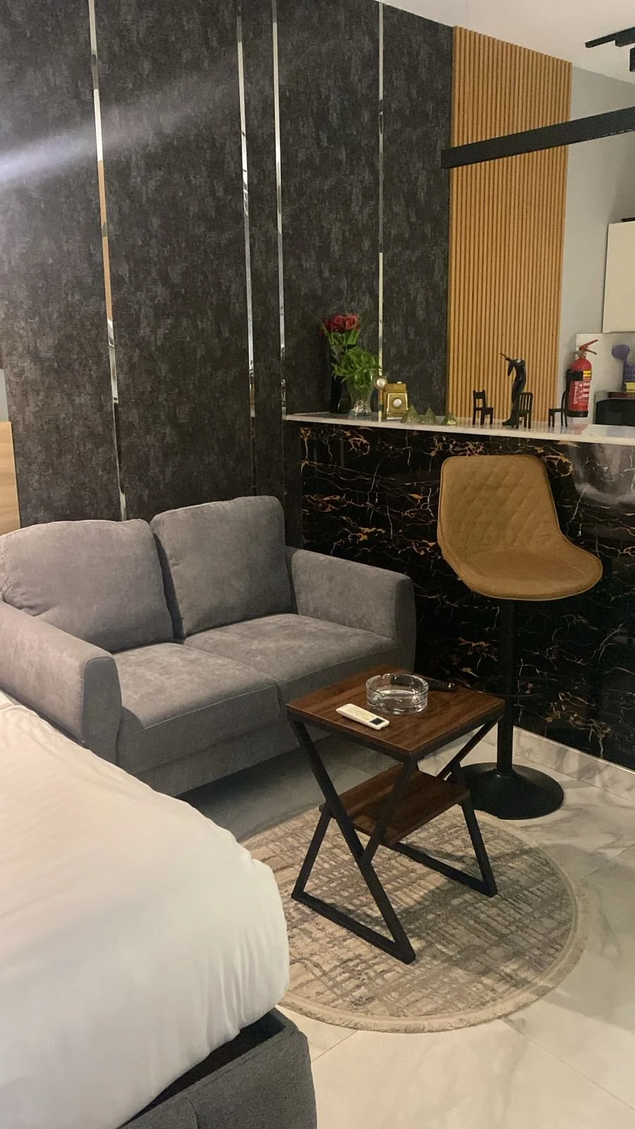 Living room with gray sofa, small wooden table with ashtray and remote, beige chair near a marble-patterned black bar with decorative items, flowers, fire extinguisher, and a black and gold backsplash, with a modern interior design.