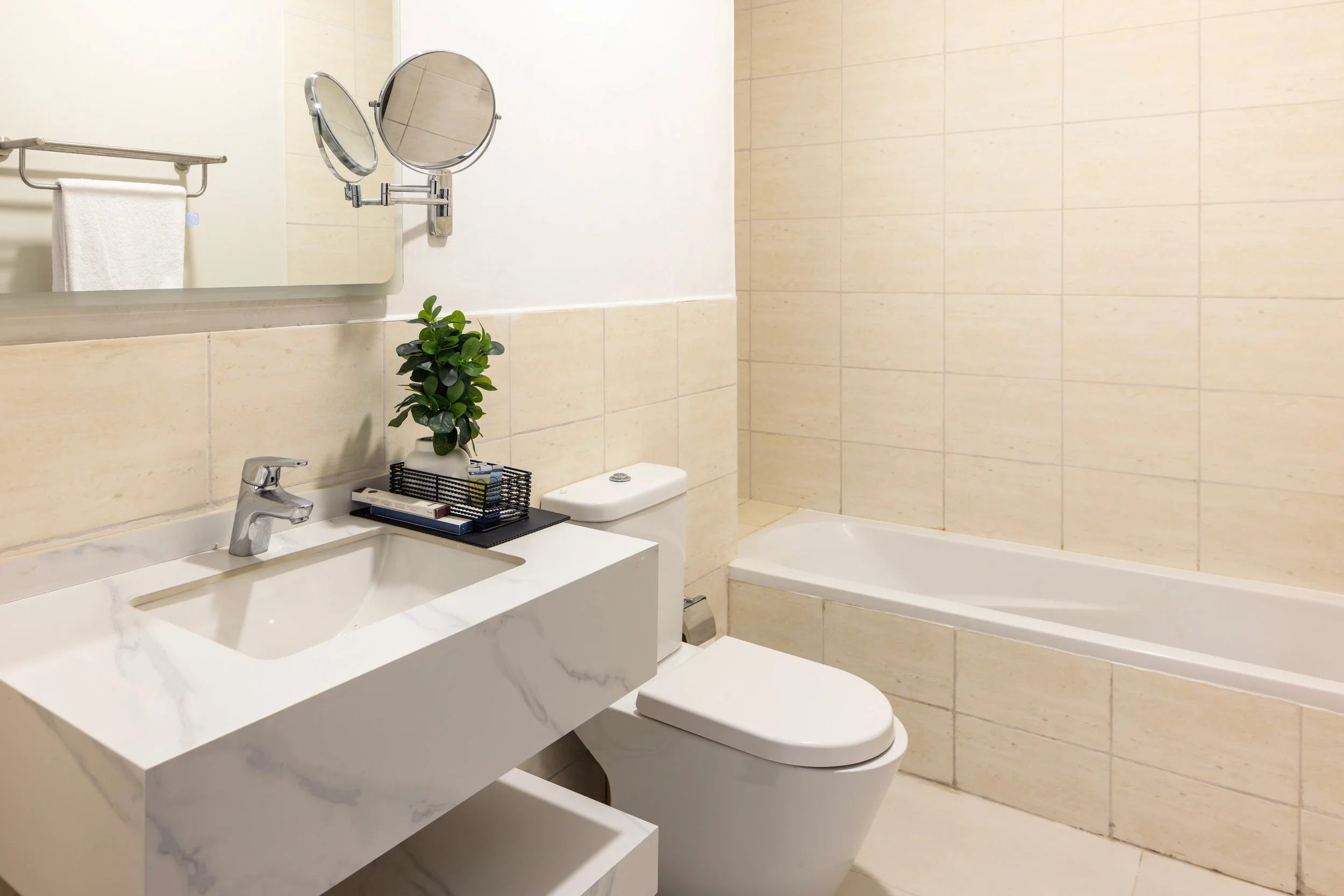 A modern bathroom with a beige tiled wall, a white toilet, a white sink with a marble pattern, a small mirror, a potted plant on the sink, a black tray with toiletries, a mirror with a magnifying side, and a bathtub.