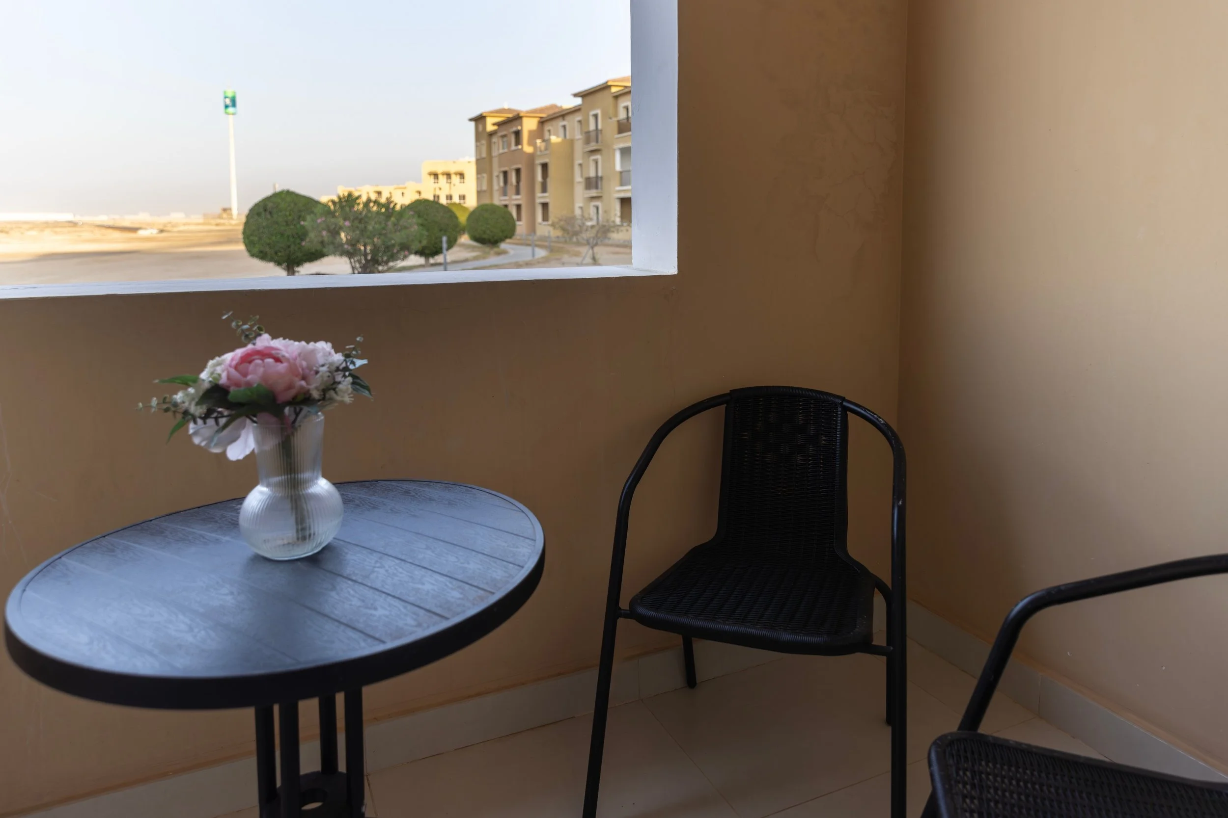 A small outdoor balcony with a black table and two black chairs, a vase with pink and white flowers, and a view of modern buildings and trees in the background.
