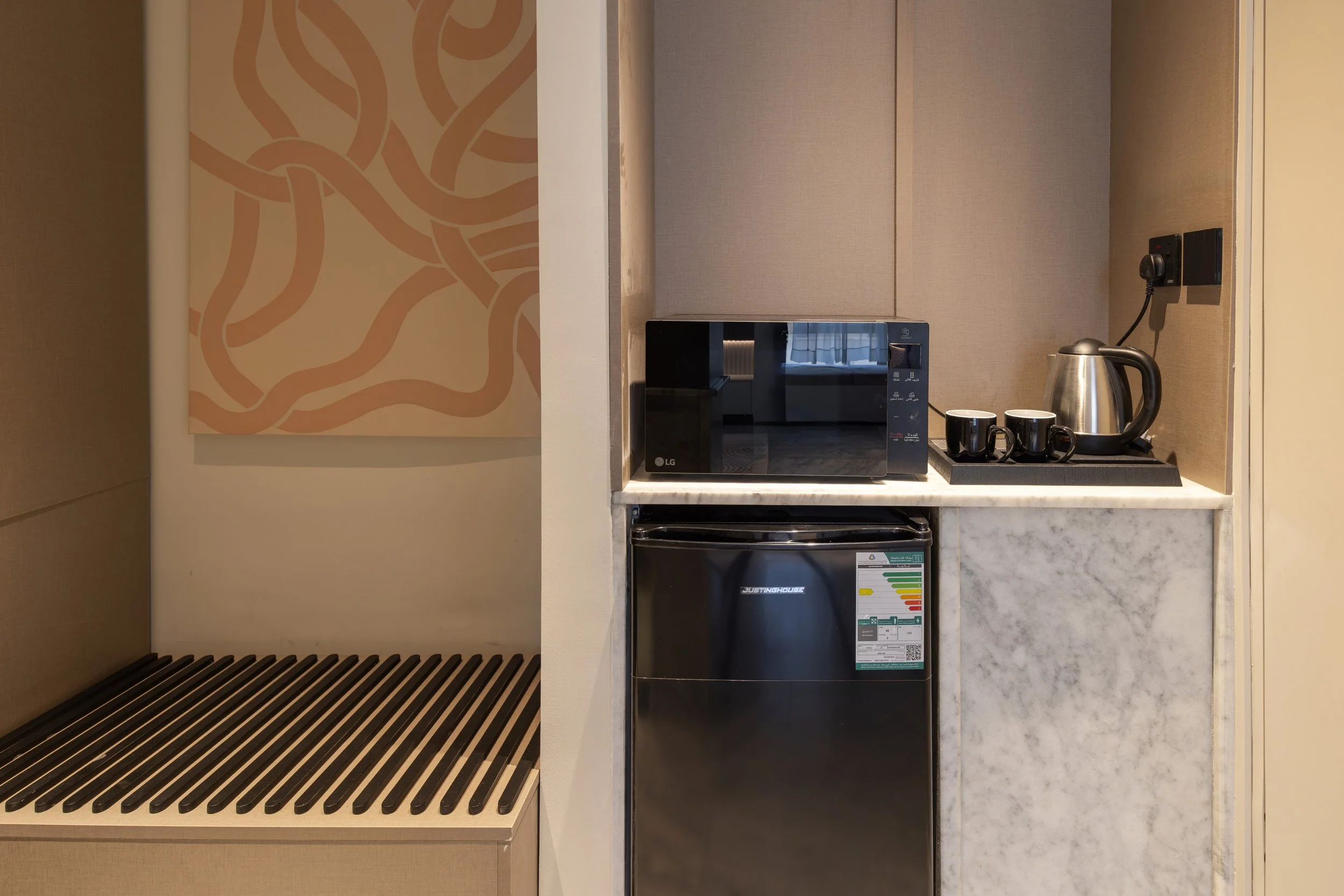 A kitchenette area with a countertop holding a microwave oven, an electric kettle, and three black cups. Below the countertop is a mini fridge with an energy efficiency label. To the left, there is a slatted bench or shelf, and on the wall, there is a piece of abstract art with swirling lines.