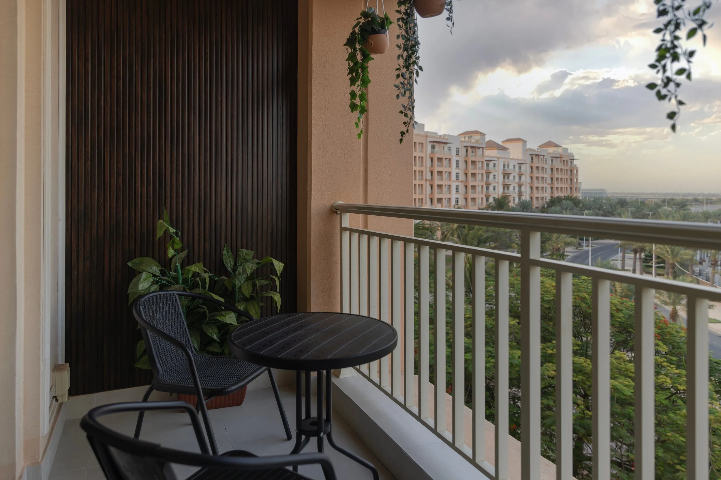 Balcony with a round table and two black chairs, potted plants, and a railing overlooking a cityscape with trees and buildings.