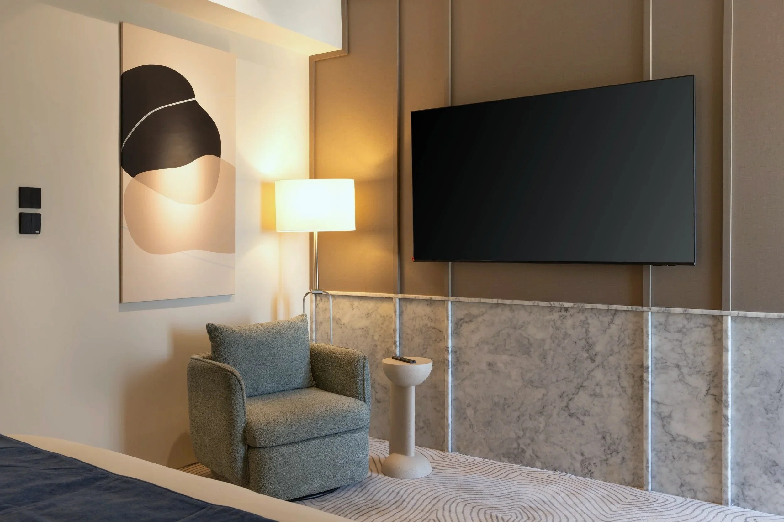 A hotel room corner with a gray armchair, a small white side table, a lit floor lamp, a large geometric abstract painting on the wall, a mounted flat-screen TV, and a partial view of a bed with a dark blue blanket.