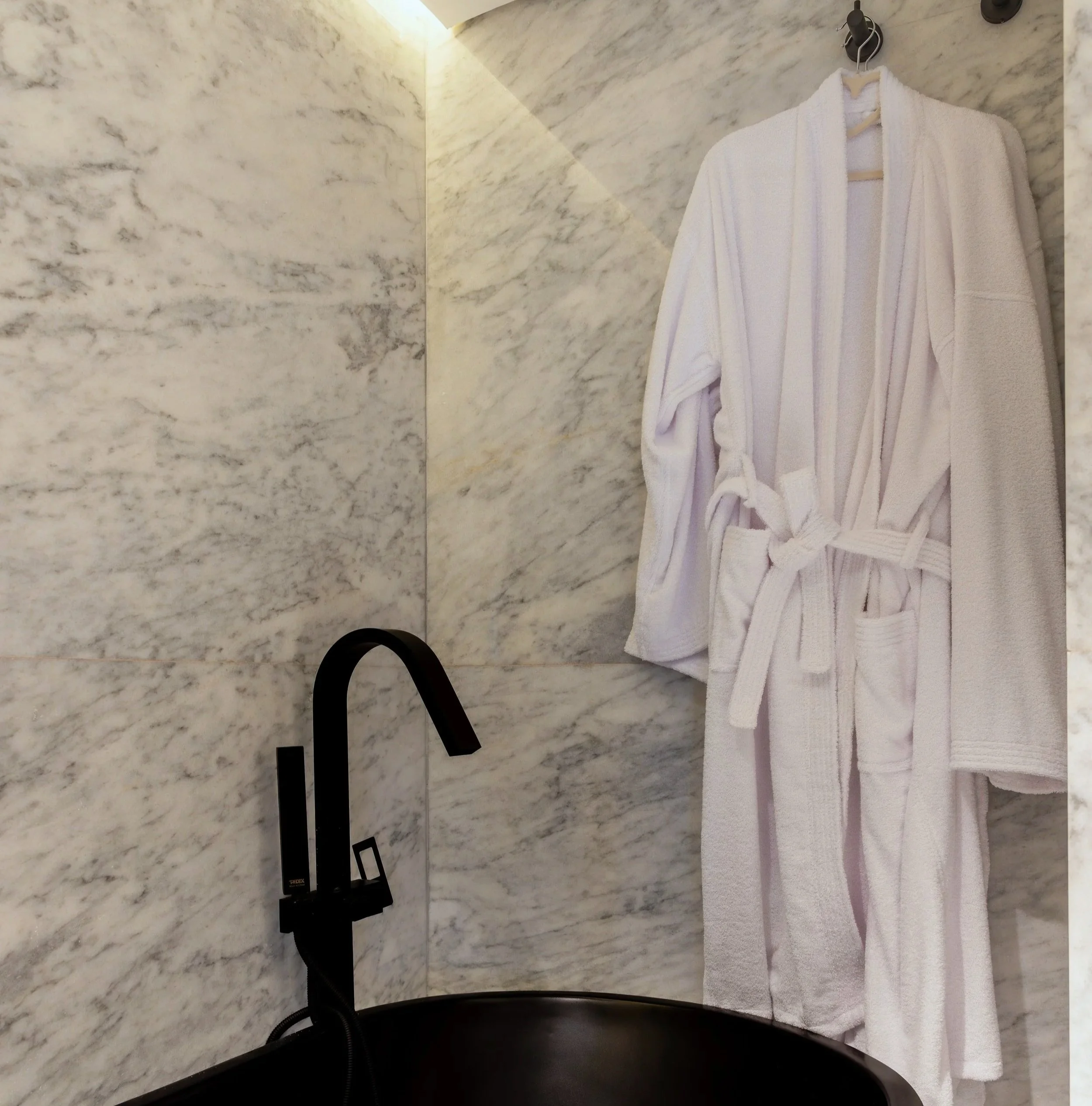 A white bathrobe hanging on a hook on a marble wall, with a black sink and faucet below.