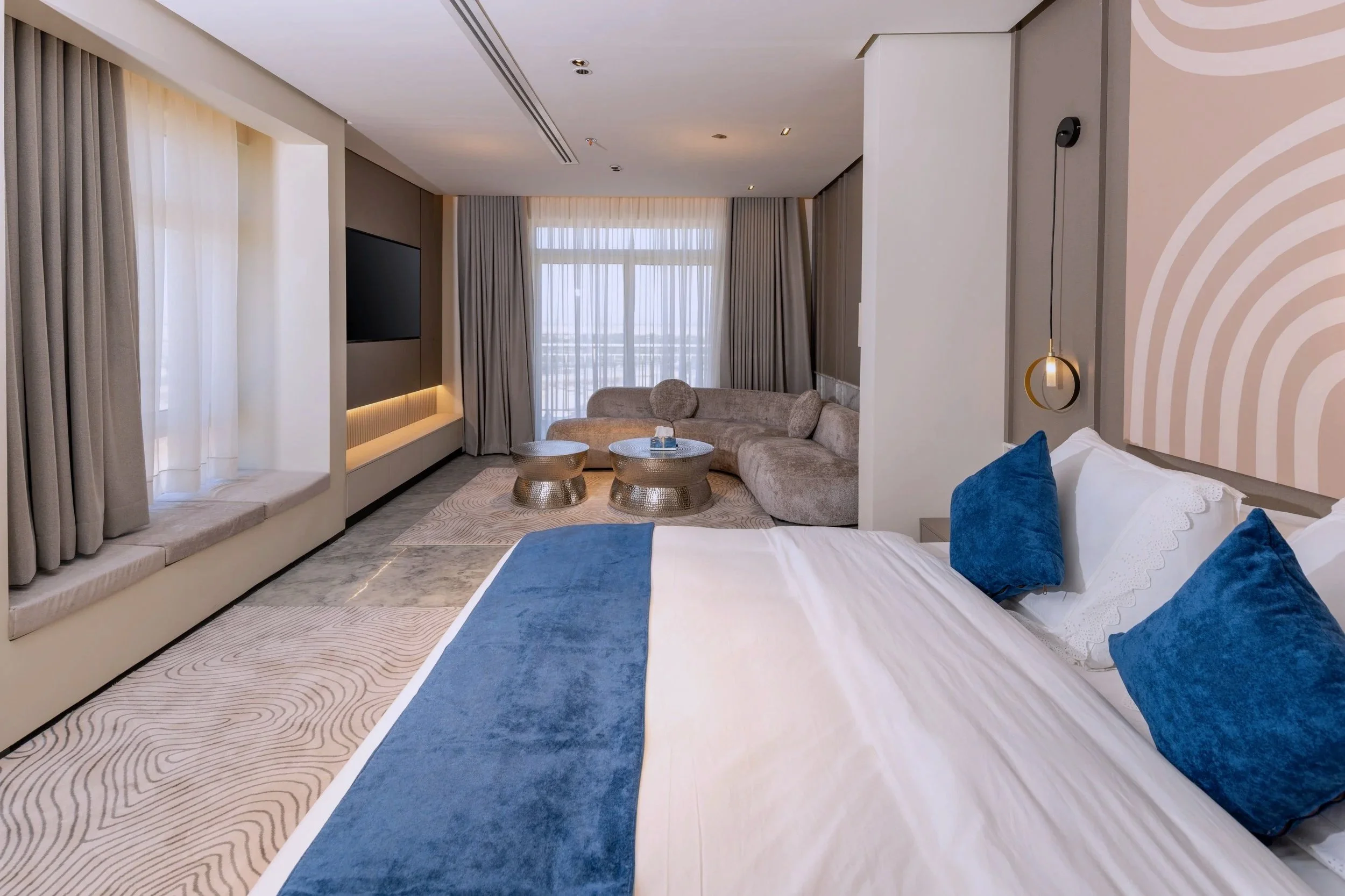 A modern hotel room with a large bed, blue pillows, sitting area with a curved beige sofa, and floor-to-ceiling windows with curtains.