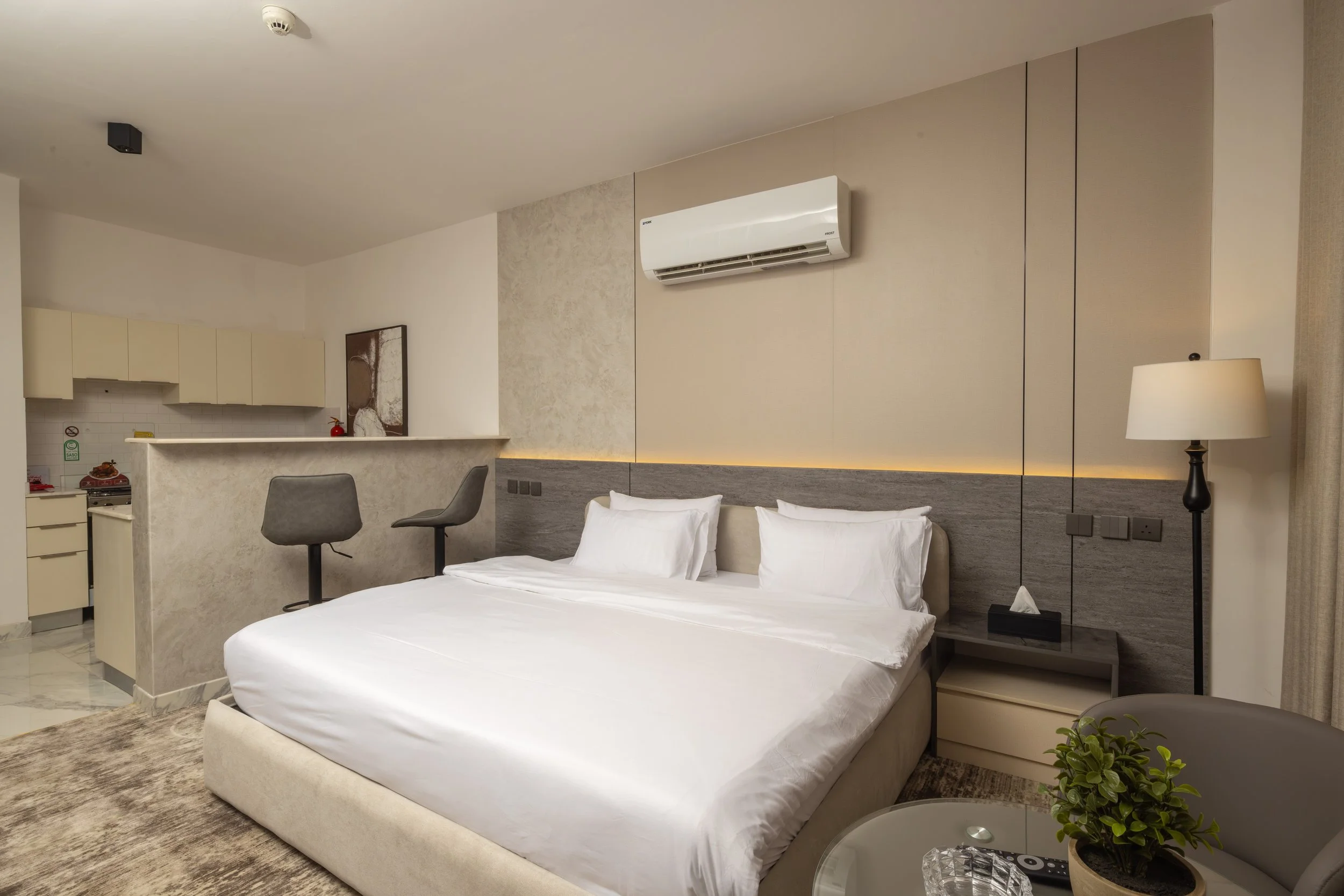 A small outdoor balcony with a black table and two black chairs, a vase with pink and white flowers, and a view of modern buildings and trees in the background.