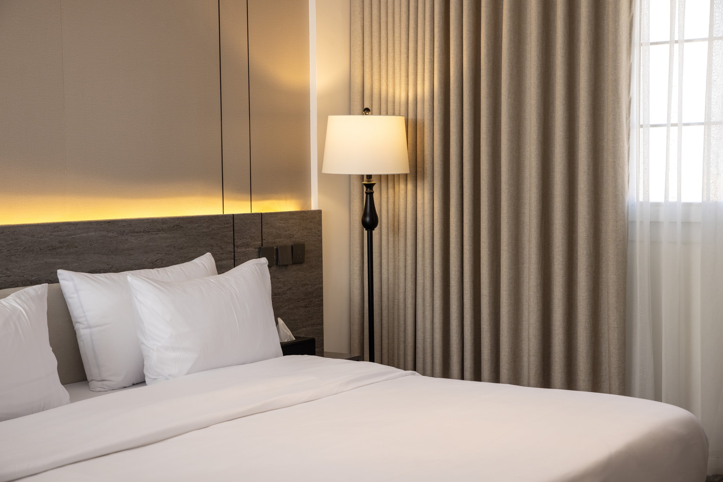 Living room with a beige sofa, brown leather chairs, a wooden coffee table, a large flat-screen TV on a wooden-paneled wall, gray curtains, a potted plant, wall art, and a modern ceiling light fixture.