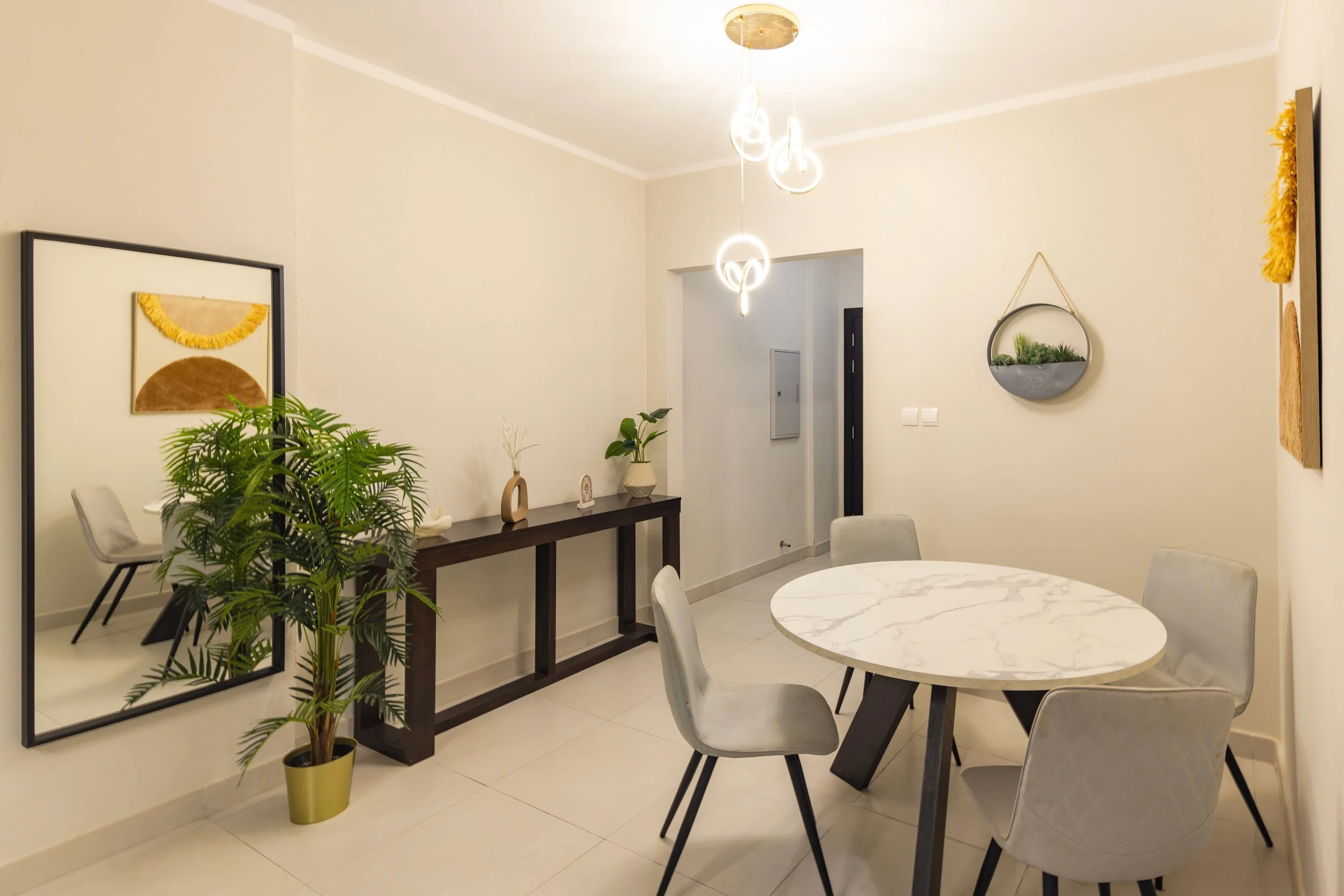 Modern dining room with a round marble table, four upholstered chairs, decorative wall art, and a console table with plants and ornaments.