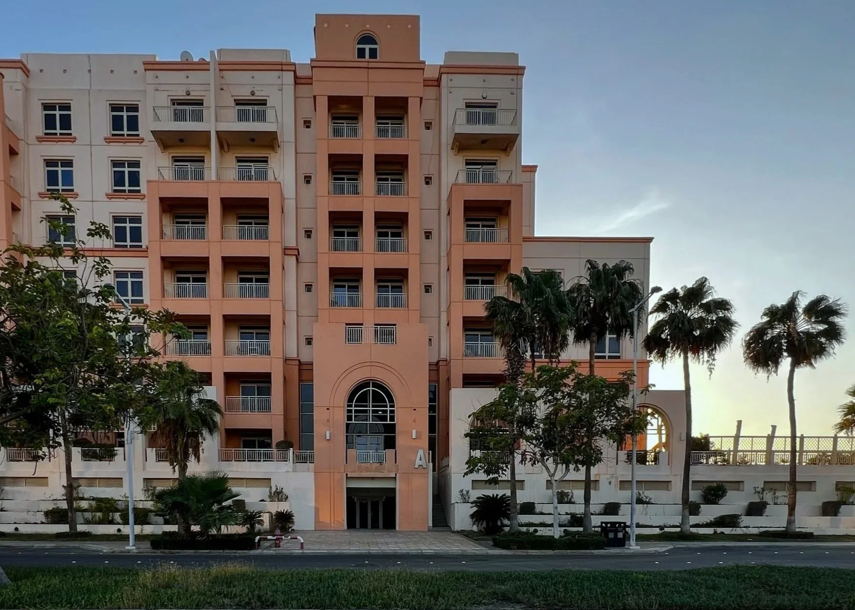 A pink and white multi-story residential building with balconies, palm trees in front, and a sidewalk with grass, under a clear sky.