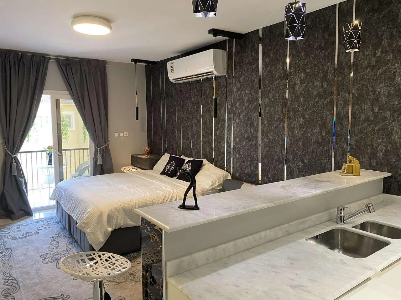 Modern studio apartment with a bedroom, black accent wall with vertical metallic strips, a bed with black and white pillows, a small balcony with outdoor furniture, and a marble kitchen counter with a sink.
