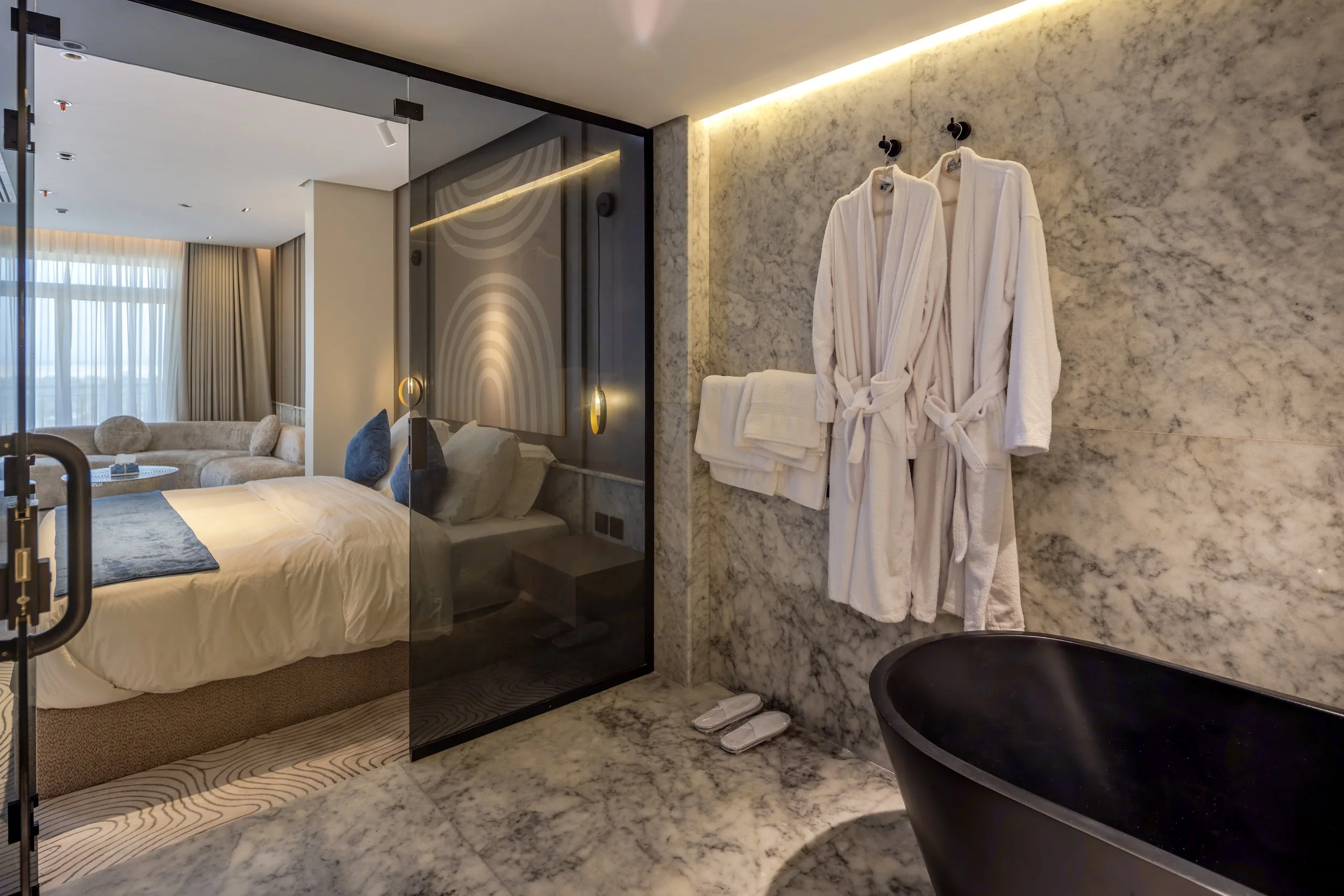 Modern hotel room with marble walls, bathrobes on hooks, slippers on the floor, and a black bathtub, separated by a glass wall from the bedroom area with a bed and seating area.