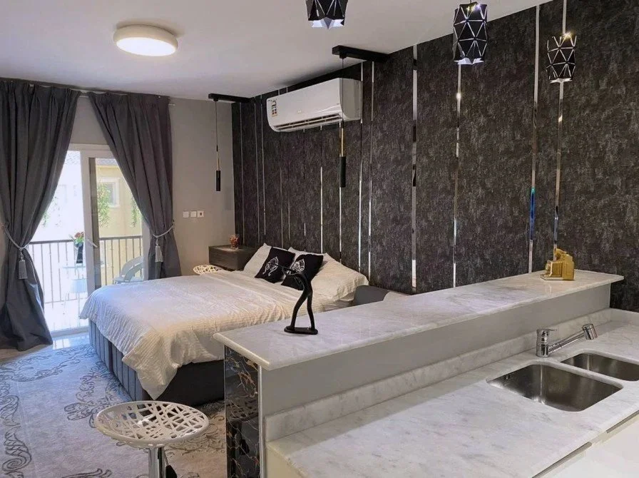 Modern bedroom with a large bed, dark accent wall, gray curtains, balcony with outdoor seating, and a countertop with a small statue and a gold phone.