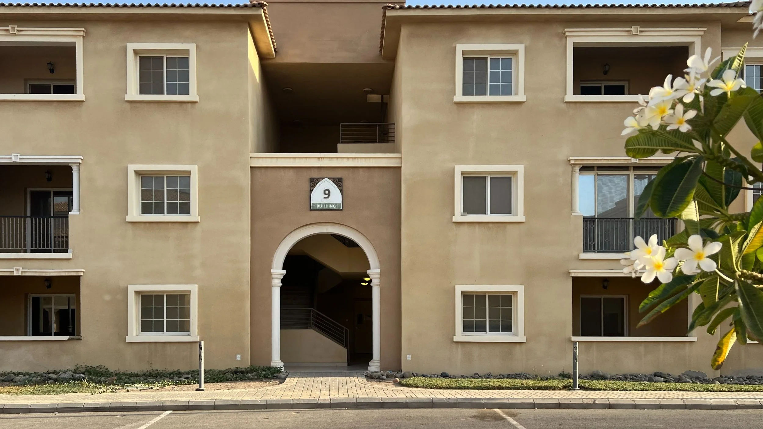 Front view of a three-story apartment building with beige walls, white window frames, and balconies, labeled as Building 9. There is a landscaped area with flowers and greenery in front, and a parking lot in the foreground.