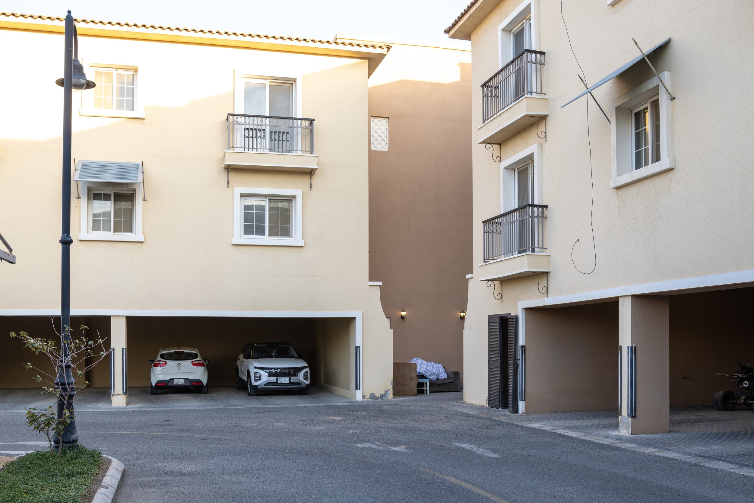 Front view of a three-story apartment building with beige walls, white window frames, and balconies, labeled as Building 9. There is a landscaped area with flowers and greenery in front, and a parking lot in the foreground.