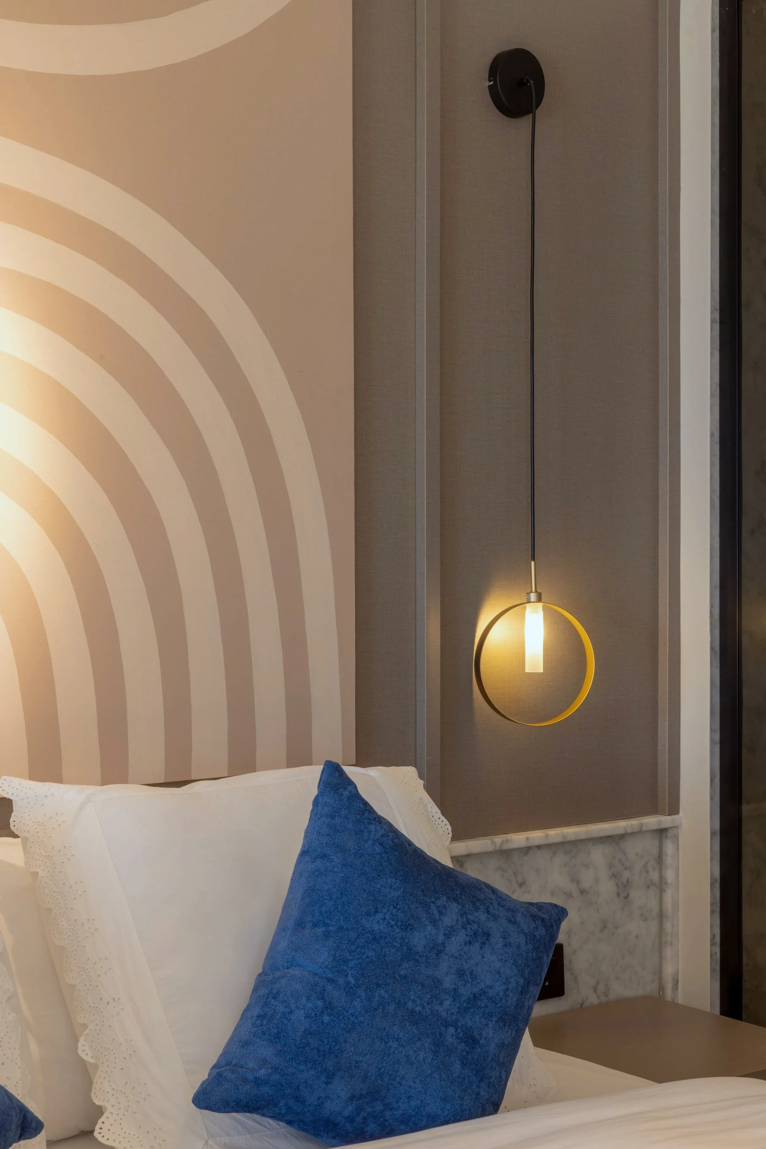 A bedroom corner featuring part of a bed with white sheets and blue pillows, a modern wall lamp with a circular design, and a wall with decorative patterns and textures.