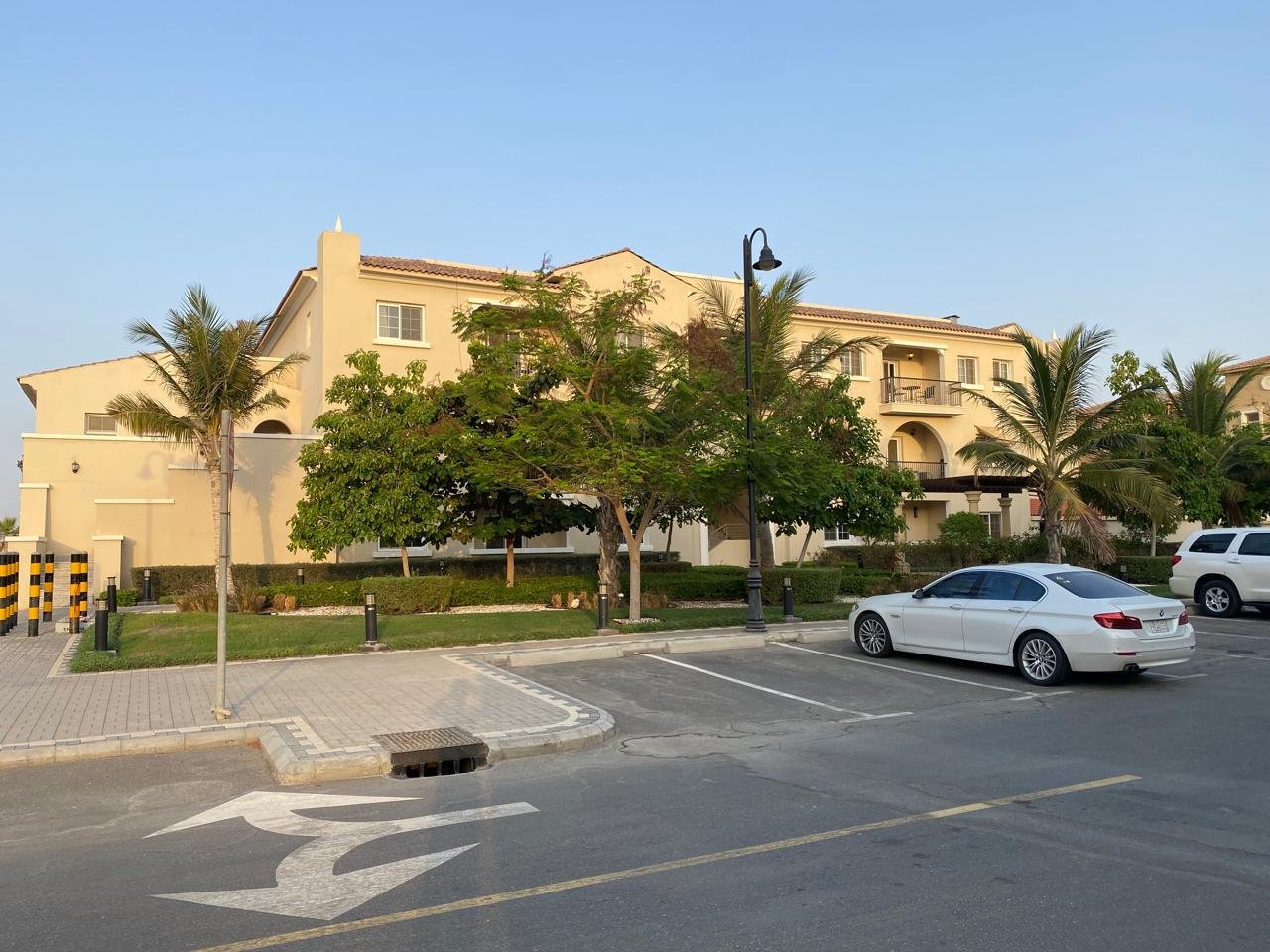 A parking lot in front of a beige apartment building with palm trees, green bushes, and a street lamp, under a clear blue sky.