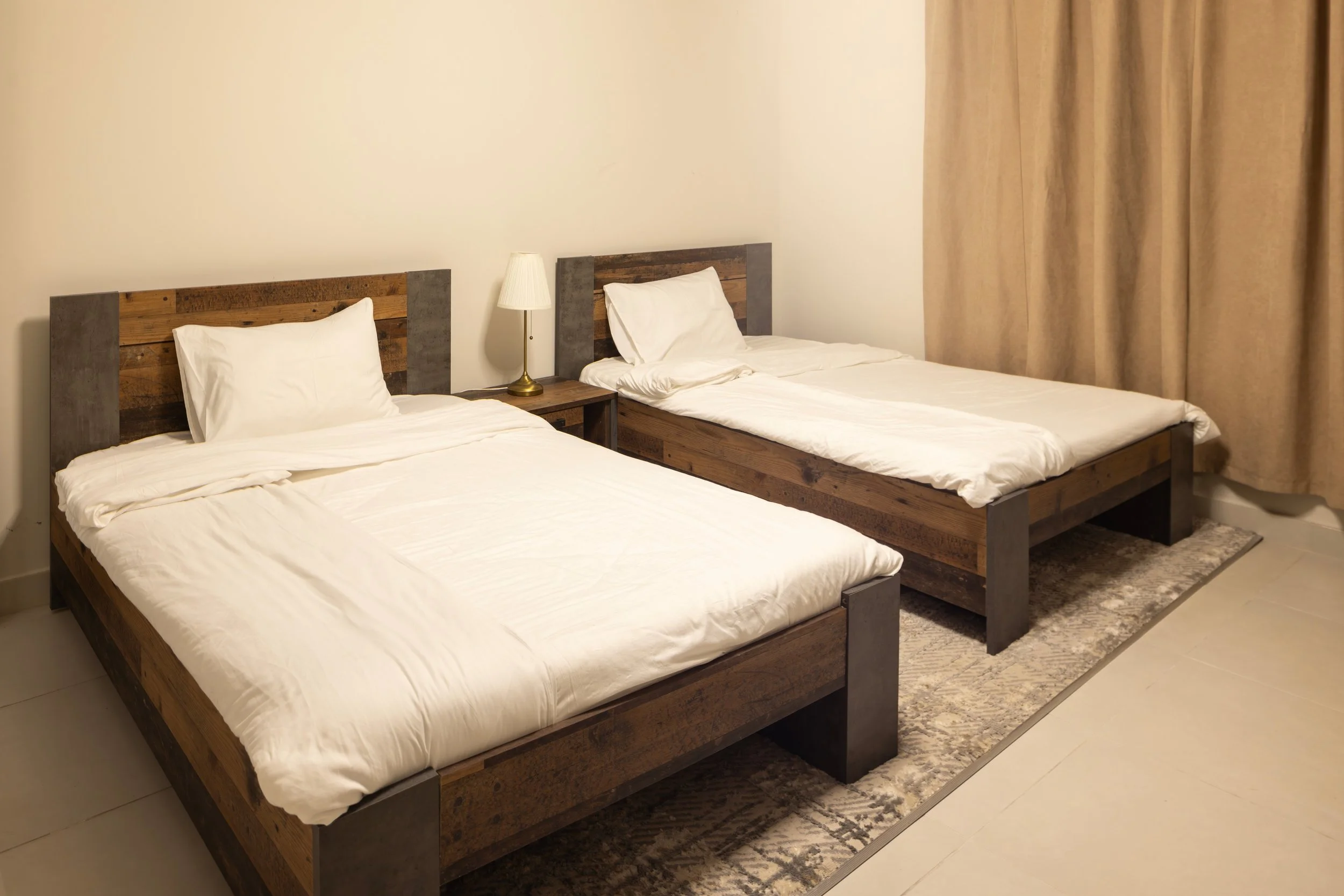 Two beds with white linens and wooden headboards, a small bedside table with a lamp, beige curtains on the side, and a rug underneath the beds.