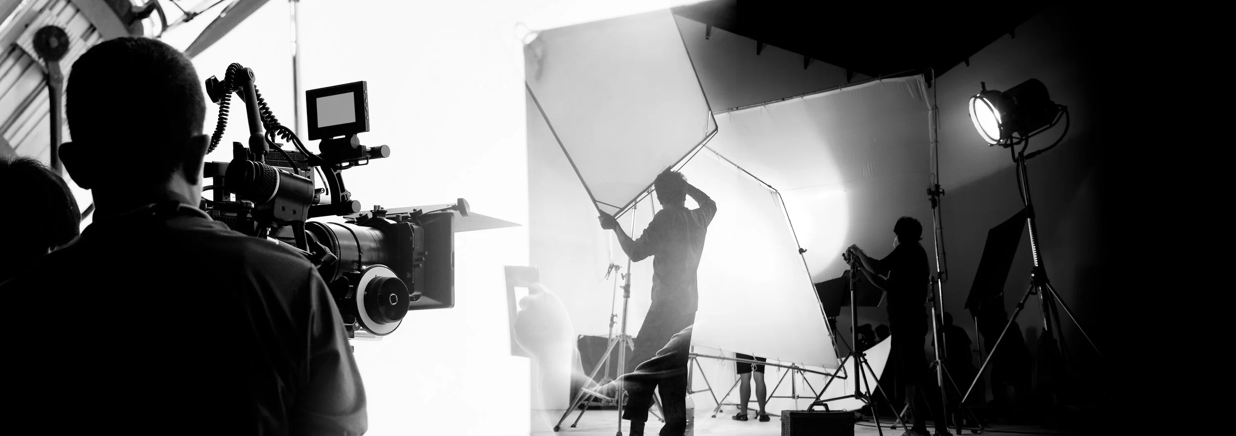 People working on a film set, with a crew member operating a camera and another person holding a large reflector, amidst studio lighting equipment and backdrop.