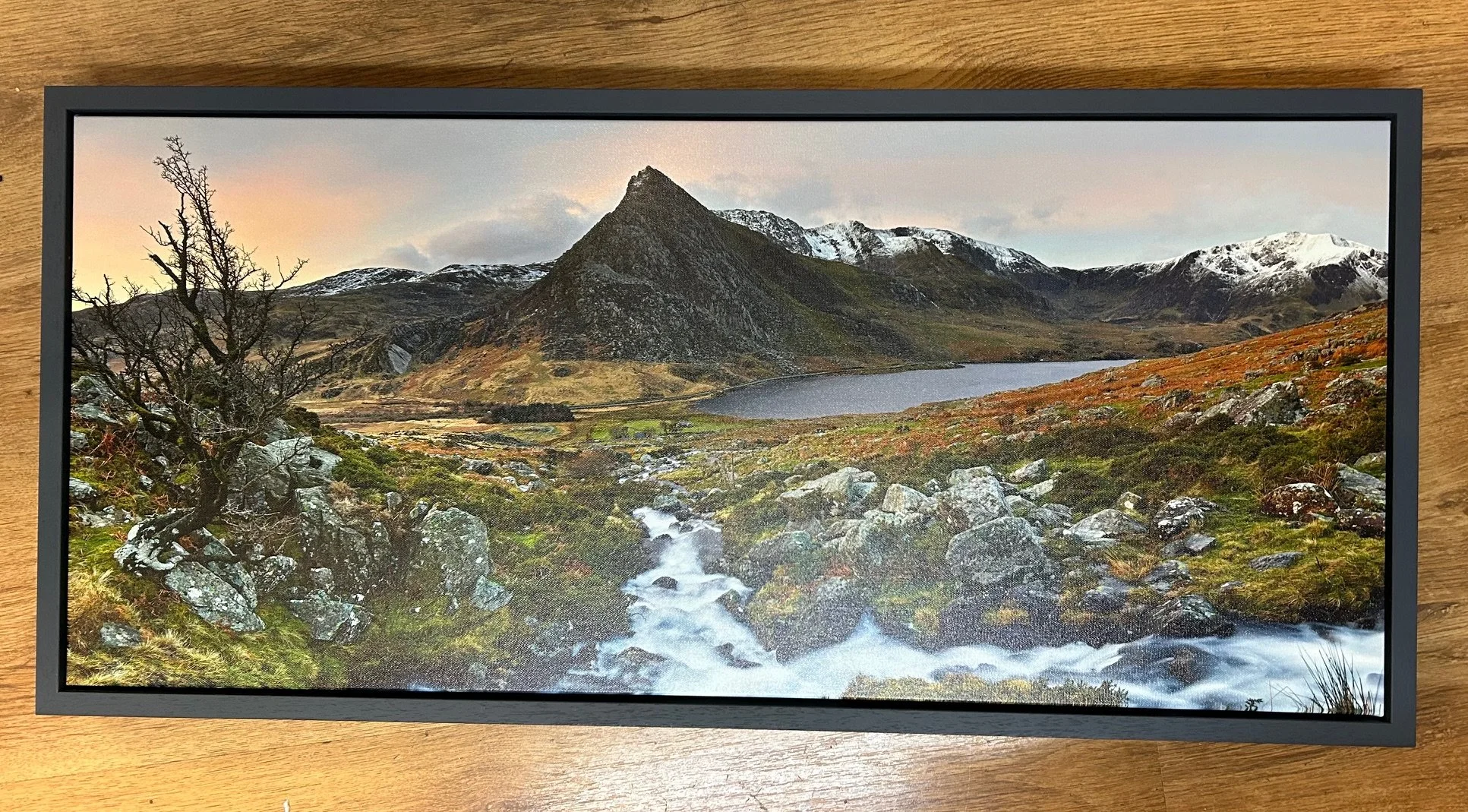 Landscape of a mountainous area with a small lake, snow-capped peaks, rocky terrain, and a stream flowing through the foreground. printed onto a canvas print and framed at not just framing