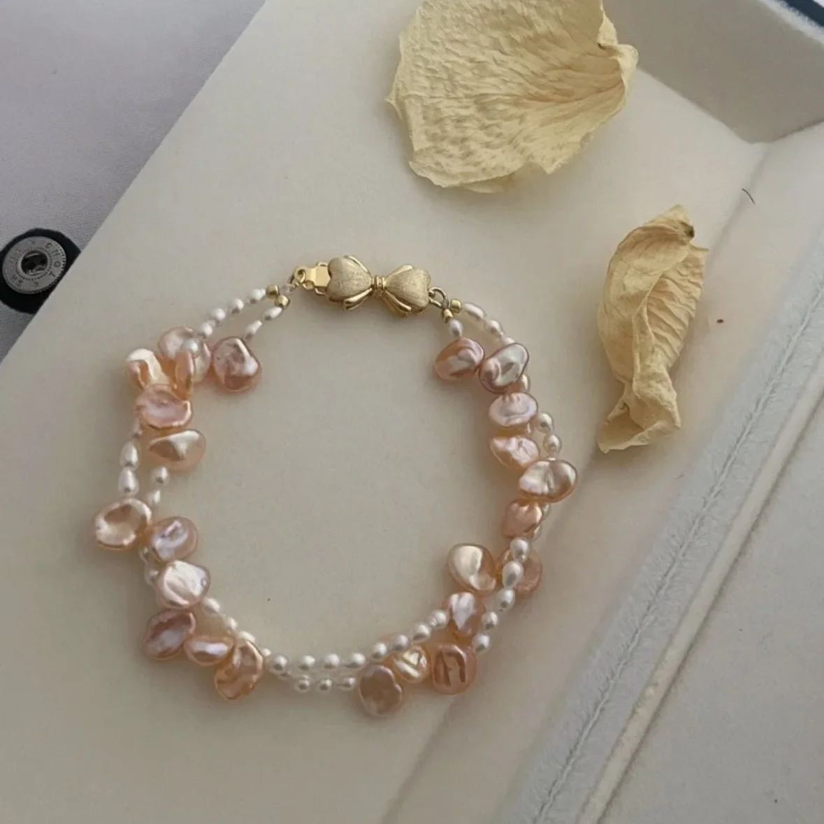 A pearl and pink shell necklace with a gold bow clasp, resting on a white surface next to two dried flower petals.