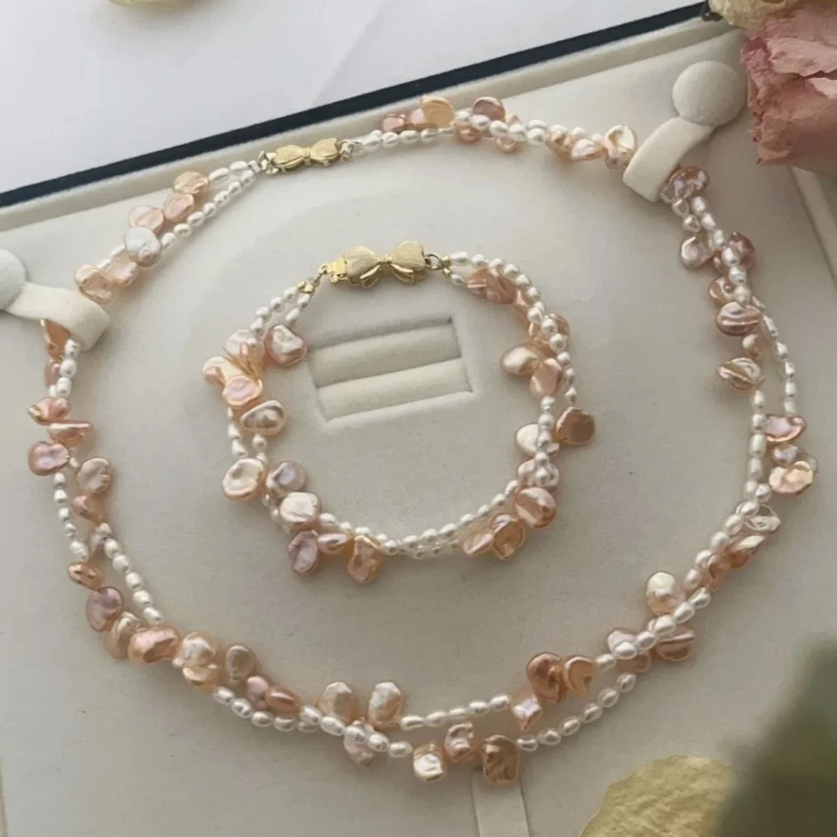 Set of pearl jewelry, including a necklace, bracelet, and earrings, arranged on a jewelry display. The jewelry features white pearls with pinkish and peachy irregularly shaped beads and gold accents.
