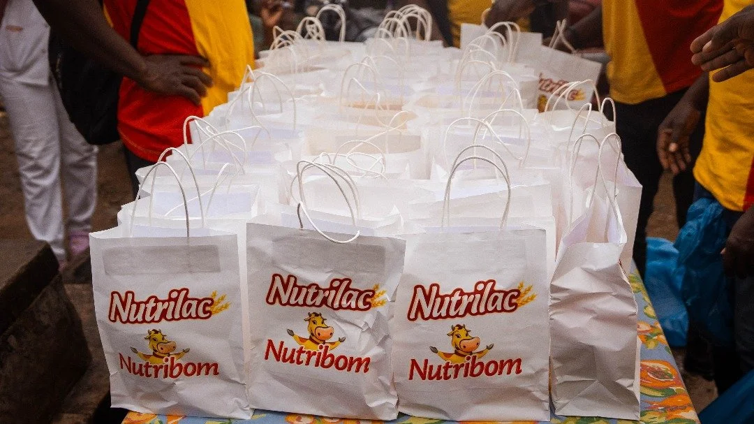 Sacs en papier blanc avec le logo Nutrilac et Nutribom, sur une table avec des personnes autour, dans un contexte de distribution ou d'événement.
