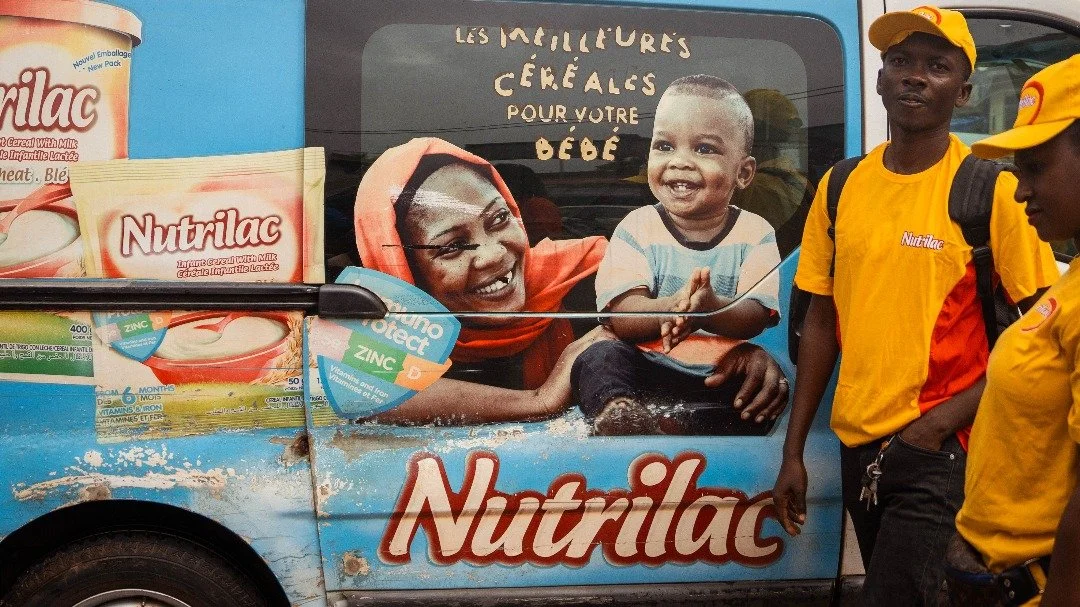 Un véhicule de promotion pour Nutrilac avec une affiche montrant une femme et un enfant souriant, entourée de personnes portant des uniformes jaunes.