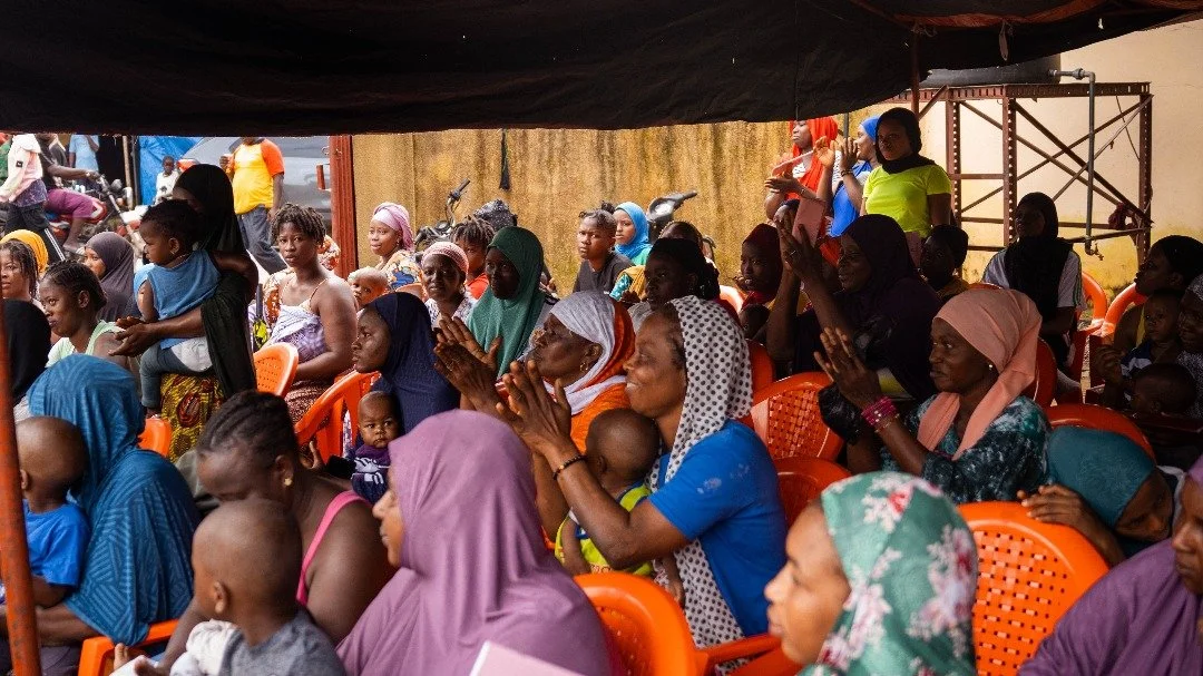Groupe de femmes et d'enfants assis sous une tente, participent à un événement communautaire, certains applaudissent, d'autres regardent attentivement.