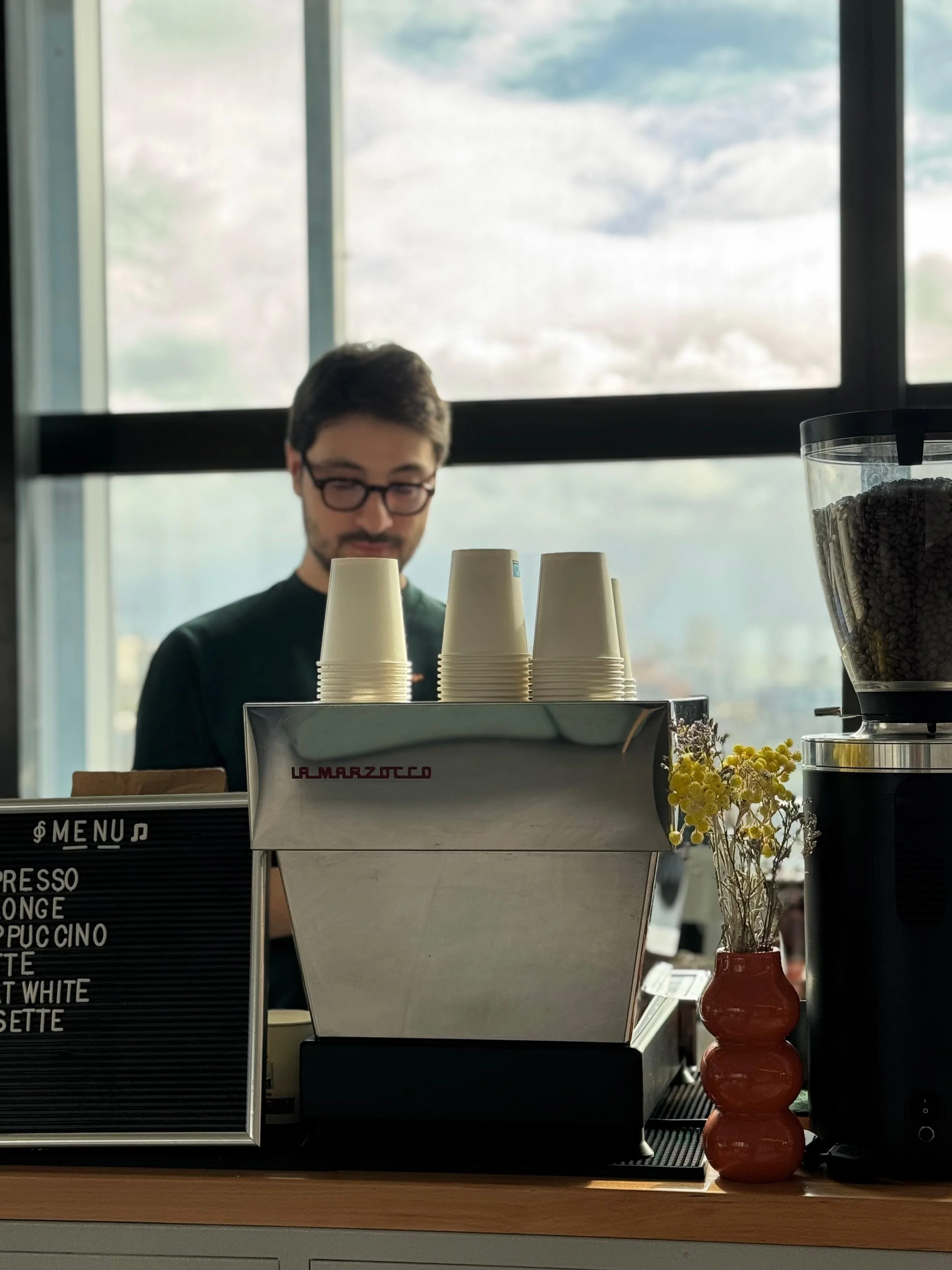 Un homme barista derrière un comptoir de café, avec des gobelets empilés et un menu en tableau noir, dans un café moderne avec grande fenêtre en arrière-plan.