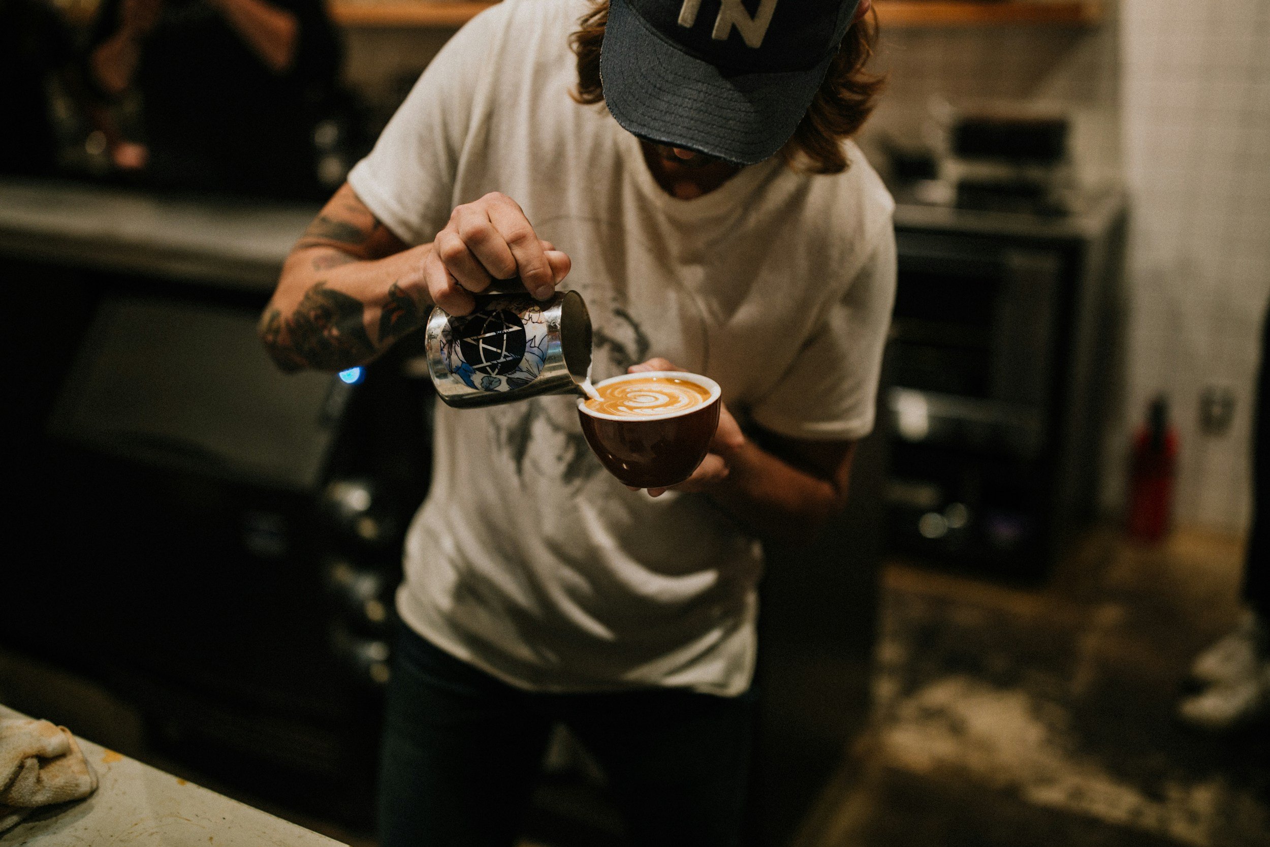 Une personne prépare un café latte avec de la crème versée dans une tasse au bar, portant une casquette noire et un t-shirt blanc, dans un café.