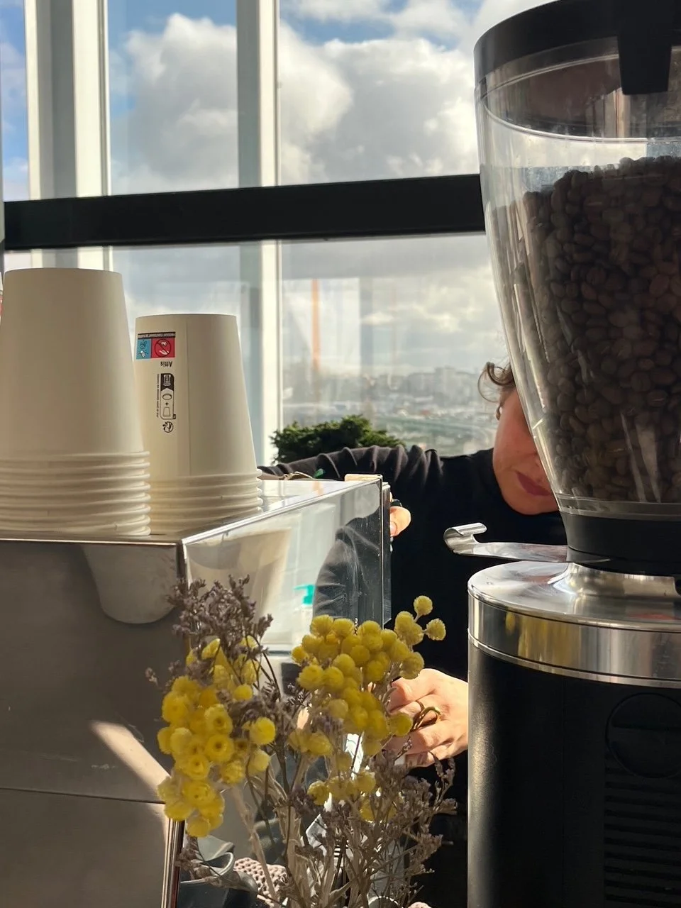 Une scène dans un café ou une station de café, avec des gobelets blancs empilés, une machine à café, un récipient de grains de café, et une personne en arrière-plan travaillant avec un grand vitrage derrière eux laissant passer une lumière naturelle 