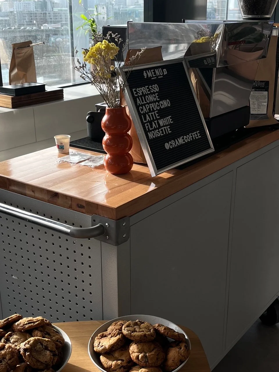 Une counter de café avec une machine à espresso, un tableau noir avec le menu, un vase avec des fleurs, deux plateaux de biscuits, et une tasse en papier.