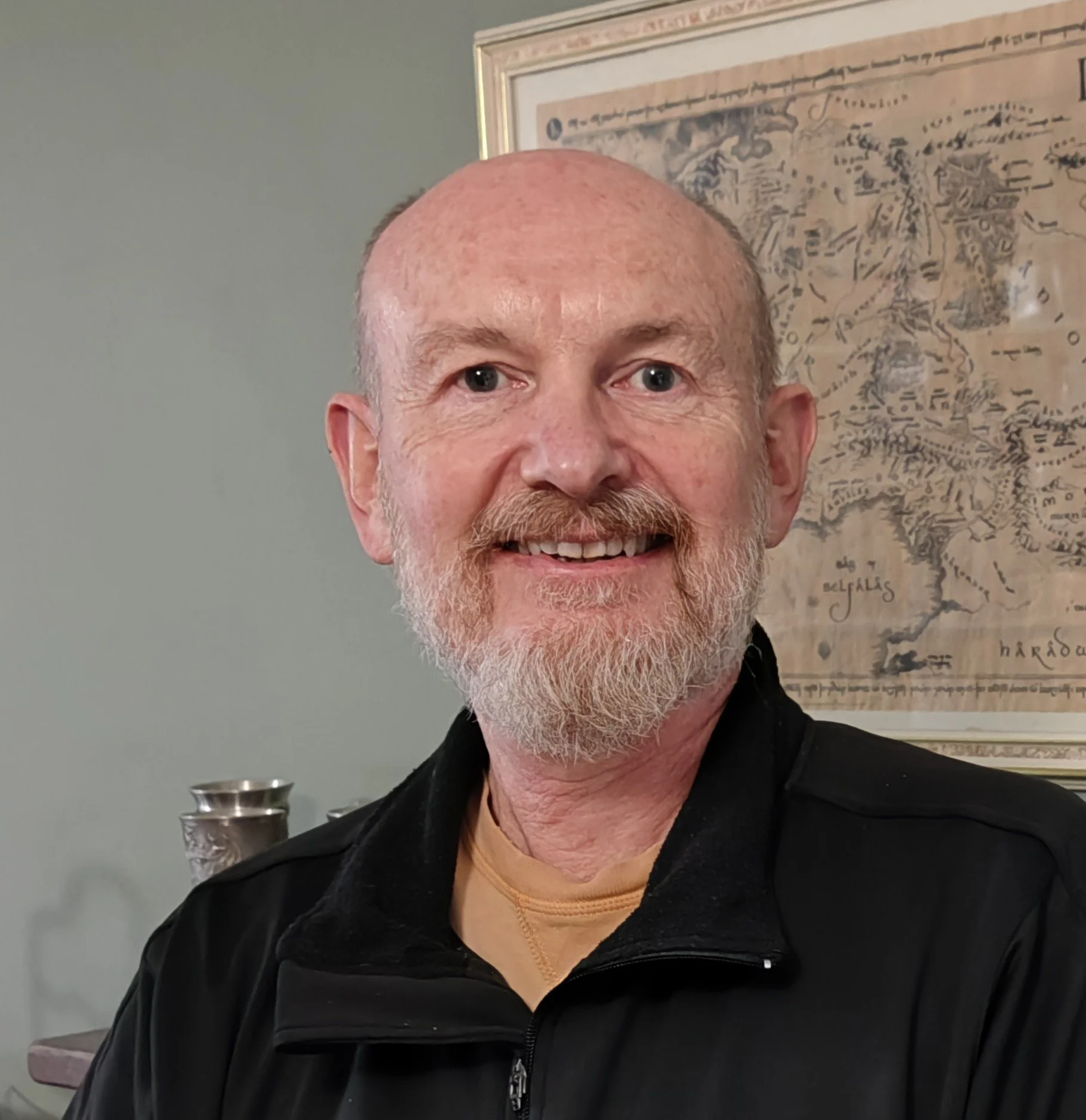 A smiling middle-aged man with a beard and bald head, wearing a black jacket, standing in front of a framed map on the wall.