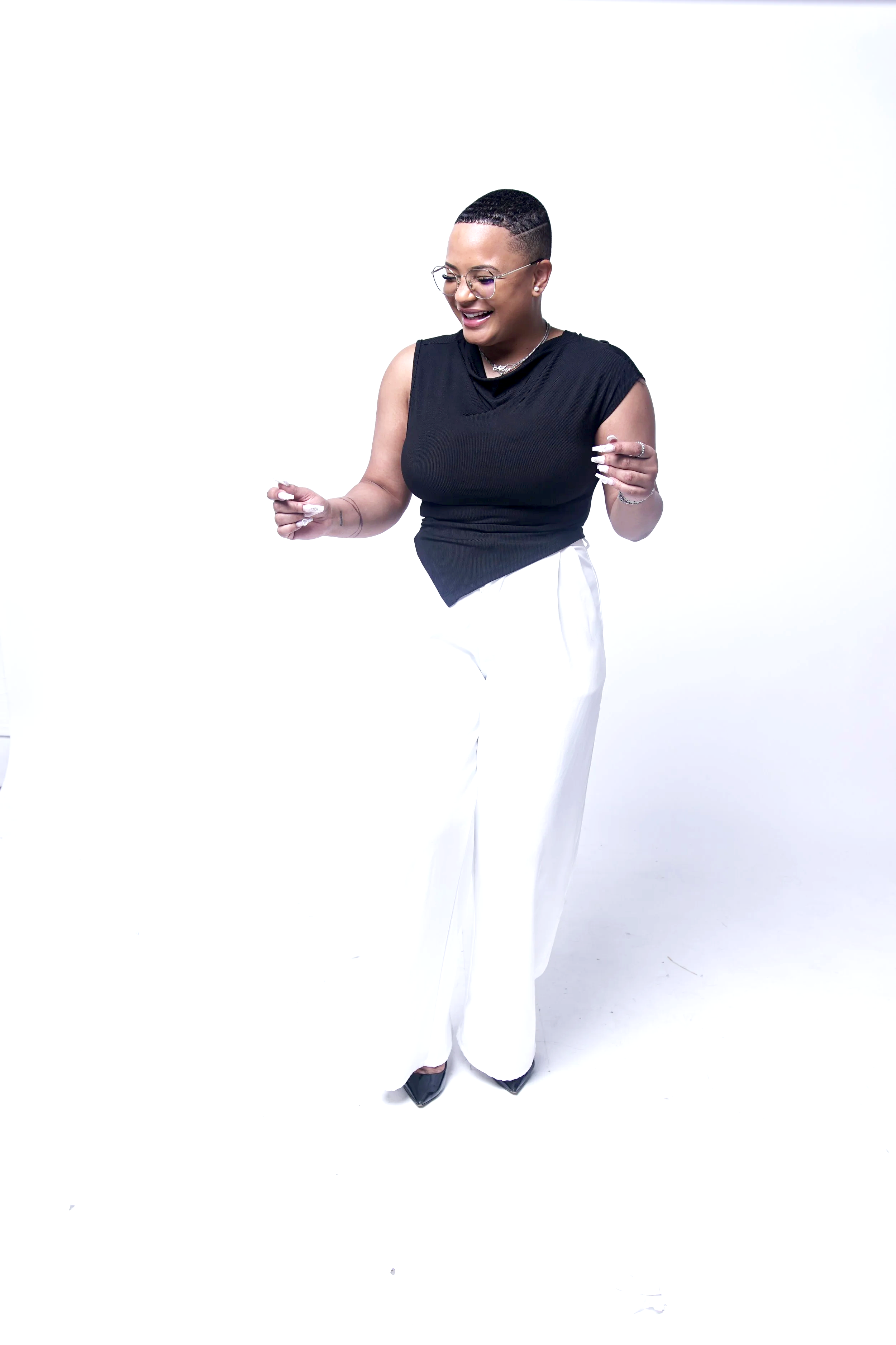 Mujer con pelo corto, gafas, top negro sin mangas, pantalones blancos de pierna ancha y zapatos negros, sonriendo y bailando sobre un fondo blanco.