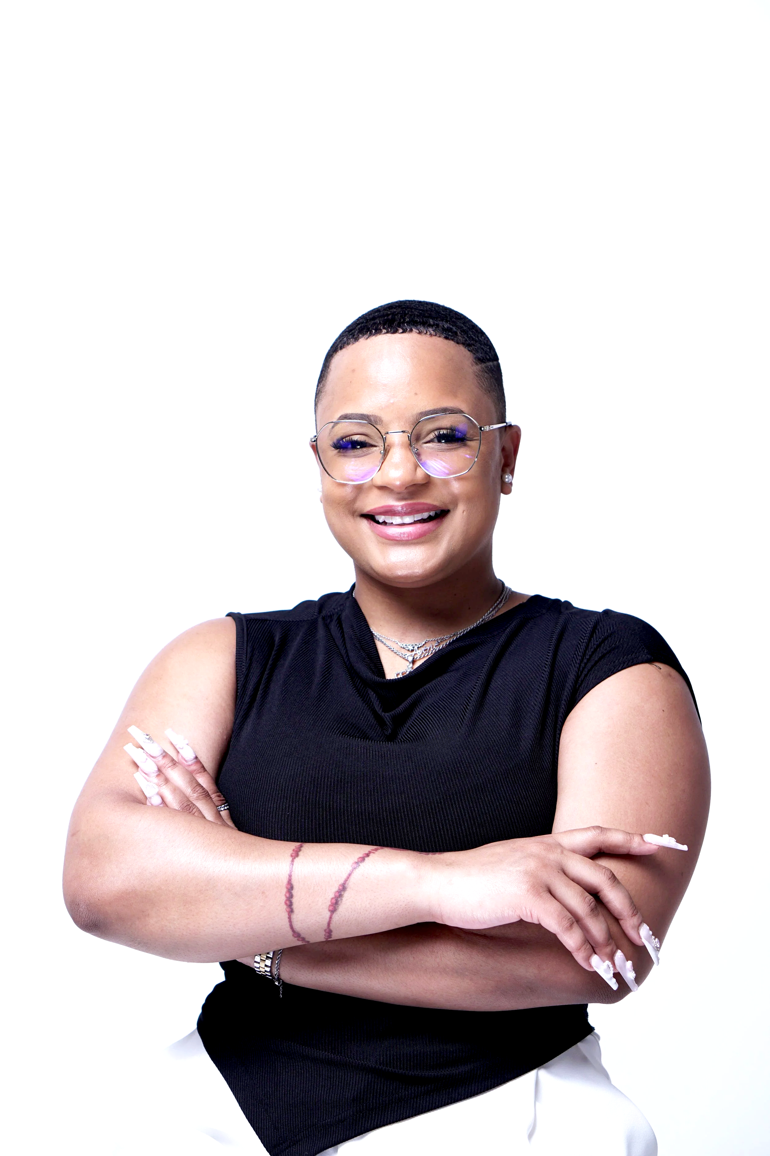 Una mujer con un corte de pelo mohicano, gafas, un top negro sin mangas y pantalones blancos, sonriendo con los brazos cruzados.