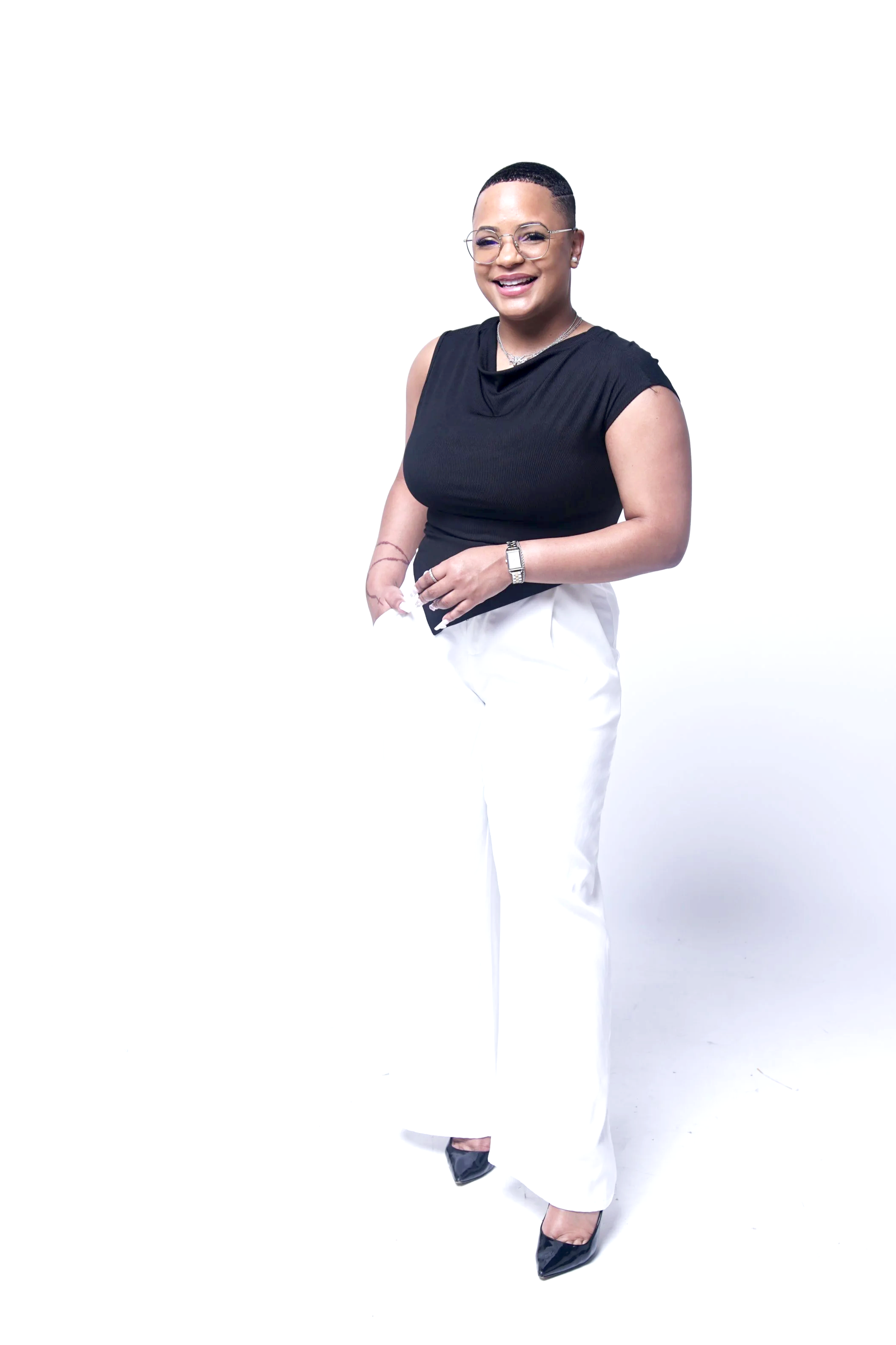 Una mujer con el pelo corto, gafas y una gran sonrisa, vestida con un top negro sin mangas, pantalones blancos, tacones negros y joyas, de pie sobre un fondo blanco.