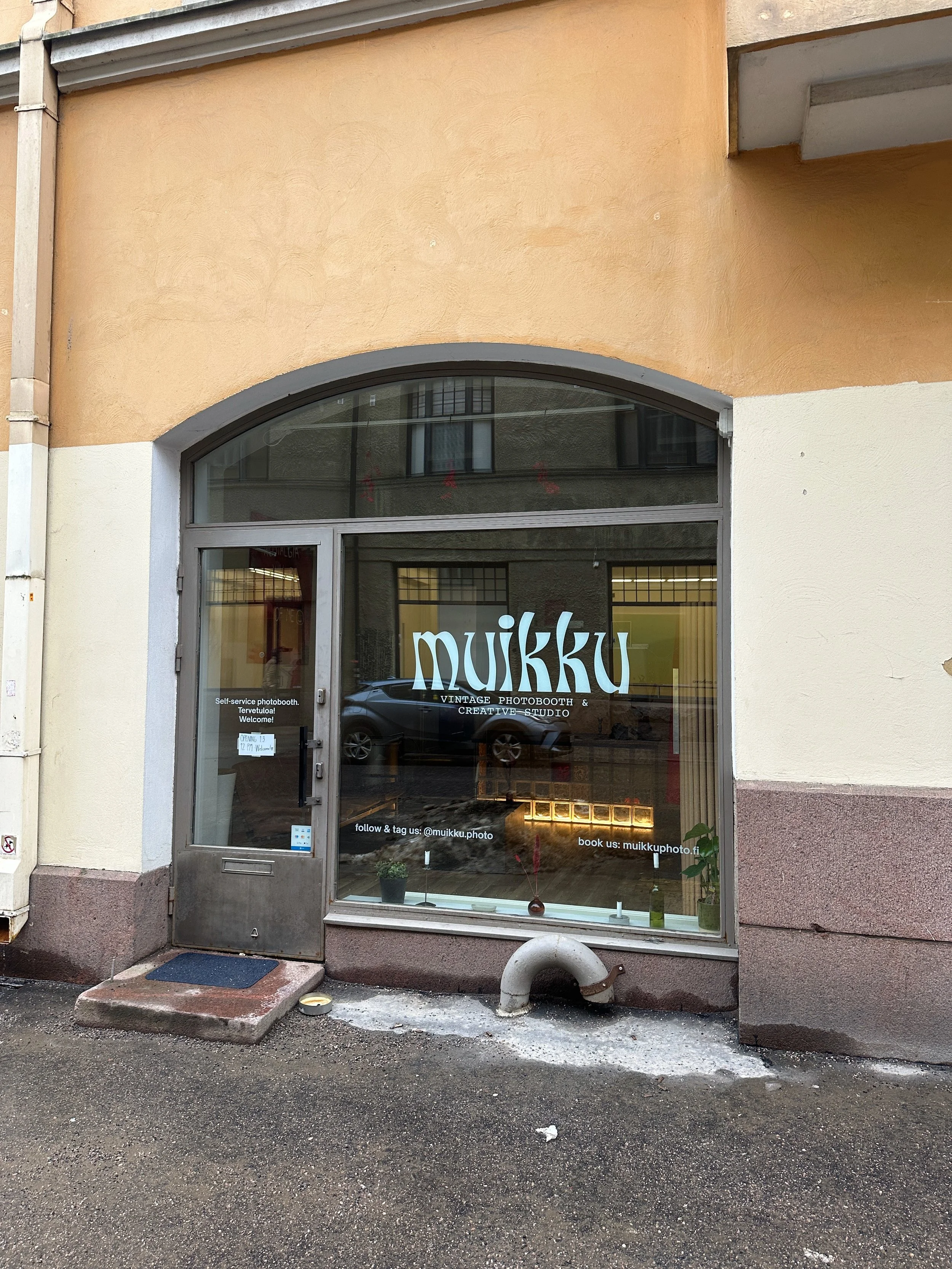 Kuvassa on valkoinen ja beige rakennus, jonka ikkunassa lukee 'muikku' ja teksti 'Vintage Photobooth & Creative Studio'. Ikkunan alla on pieni sisäänkäynti ja ikkunalaudalla on pieniä kasveja.
