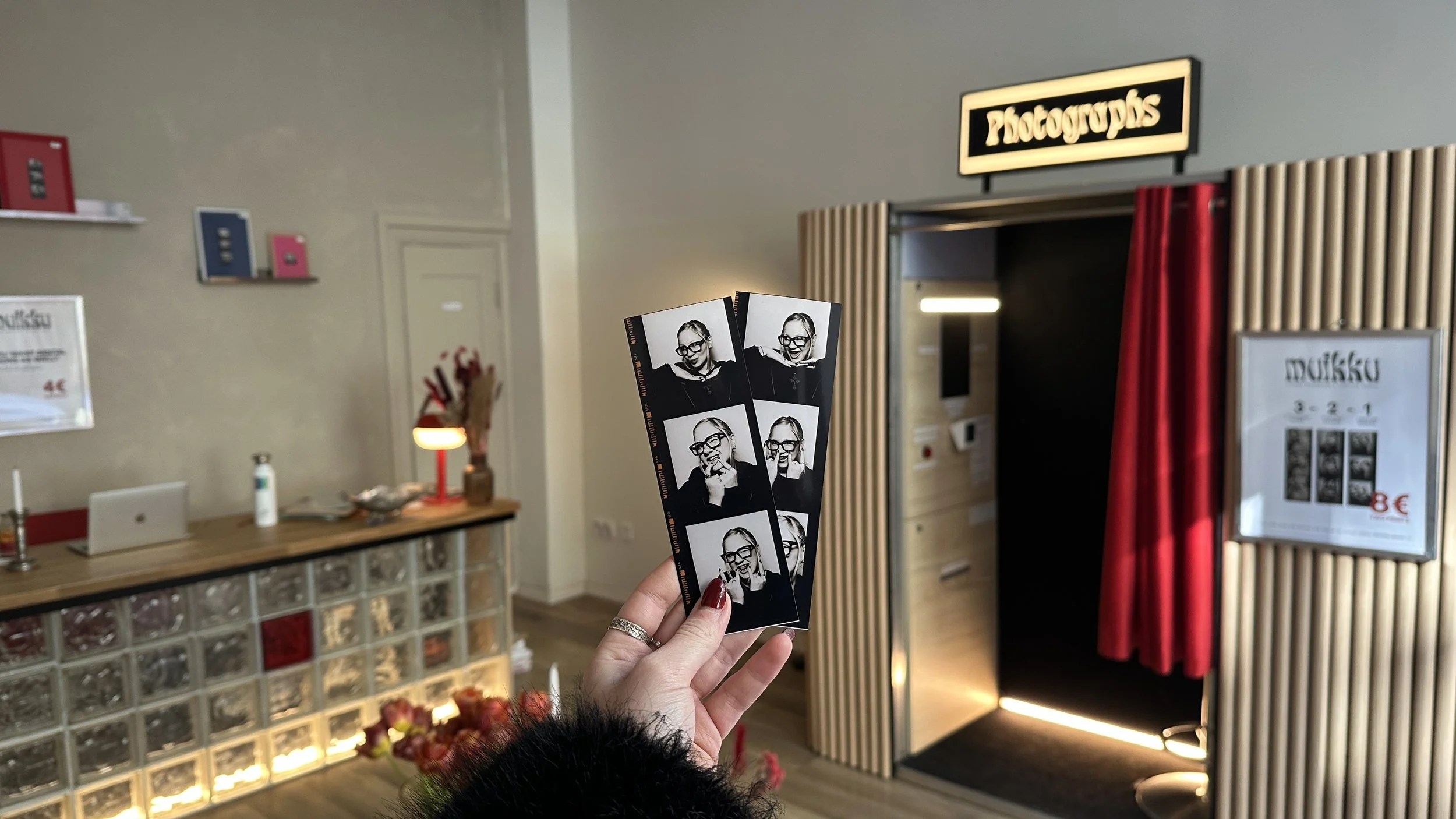 Henkilö pitää kädessä valokuvia valokuvapisteessä, jossa on valokuvapisteen merkki ja verho taustalla. Välillä on pieni pöytä ja rekvisiittalaite.