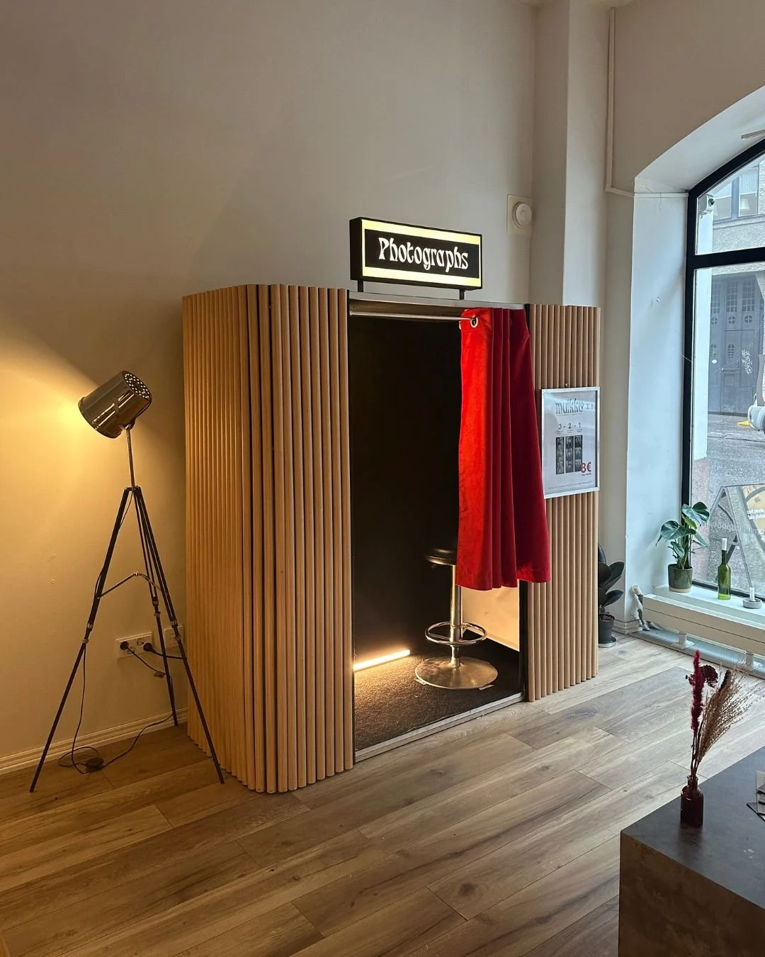 Valokuva kahvahuoneesta, jossa on valaisintason ja jalustavalaistuksen lisäksi pieni valkoinen valomainos, jossa lukee 'Photographs', sekä keltainen verho, joka peittää valokuvaajan ständin. Huoneessa on puulattia ja suuri ikkuna, jossa näkyvät rakennukset ja pieniä huonekasveja ikkunalaudalla.