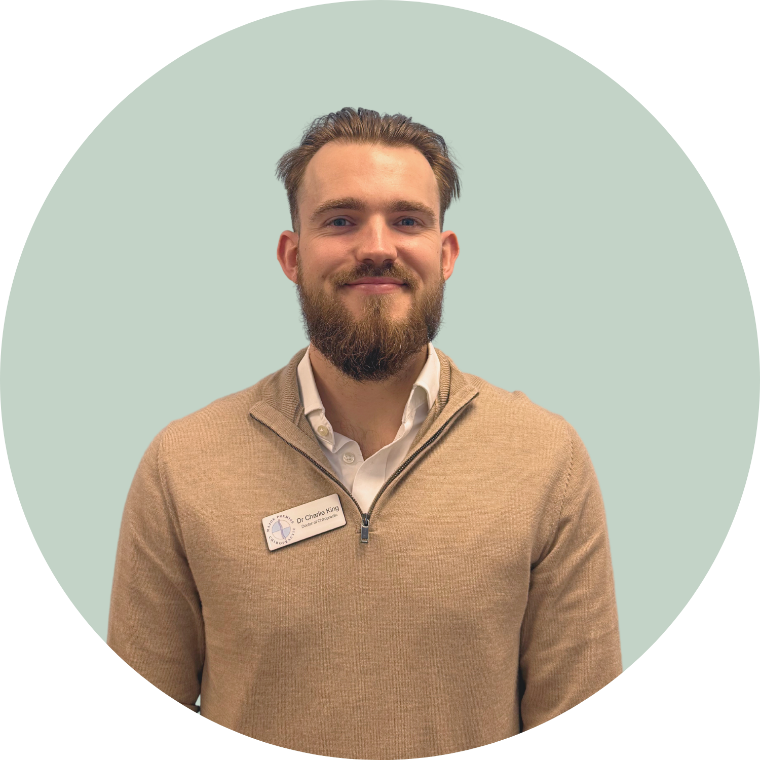 Portrait of a smiling man with a beard wearing a beige zip-up sweater and white use, standing against a light green background.