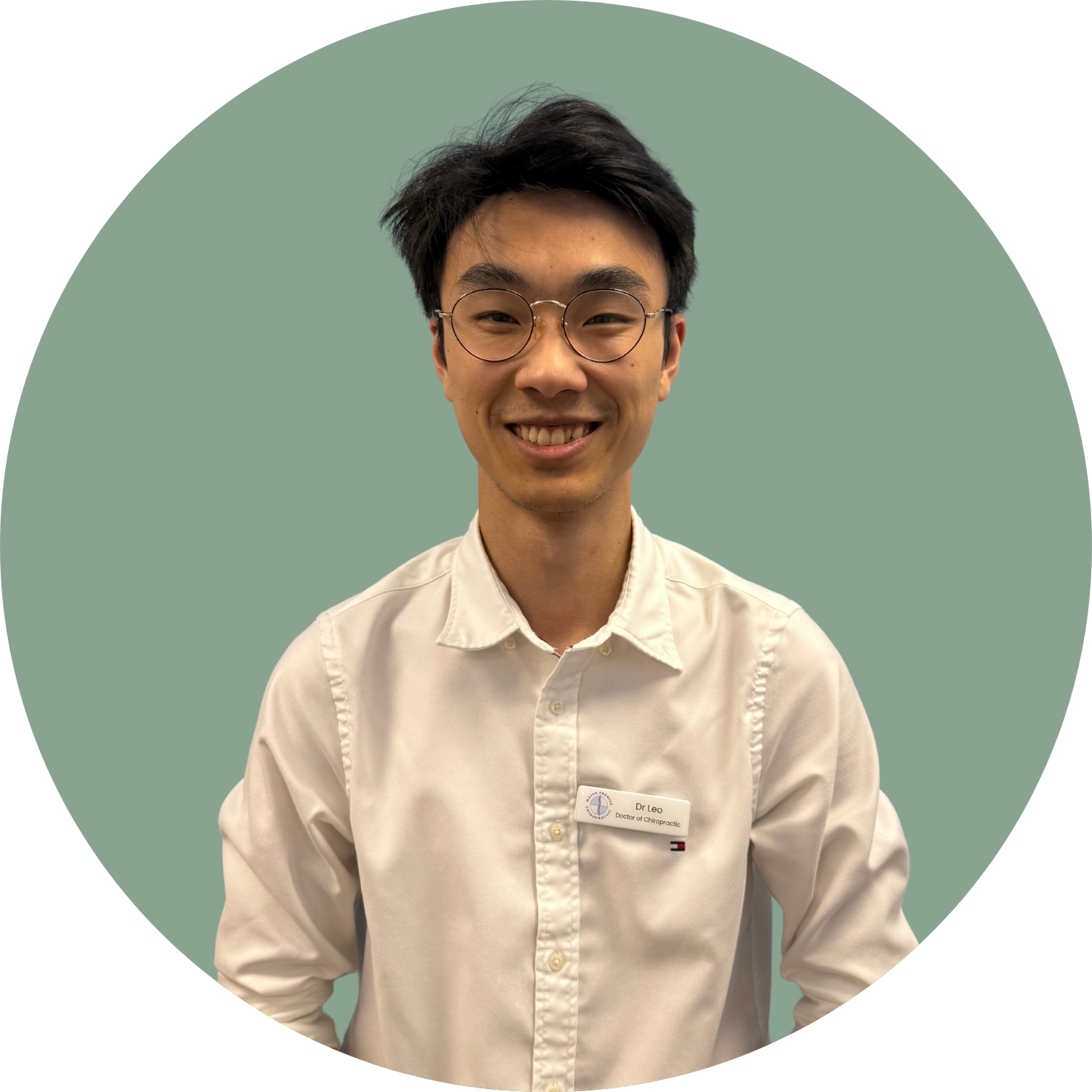 A young man smiling wearing round glasses and a white shirt. He has a name tag that says Dr Leo, Doctor of Chiropractic, against a muted green background.