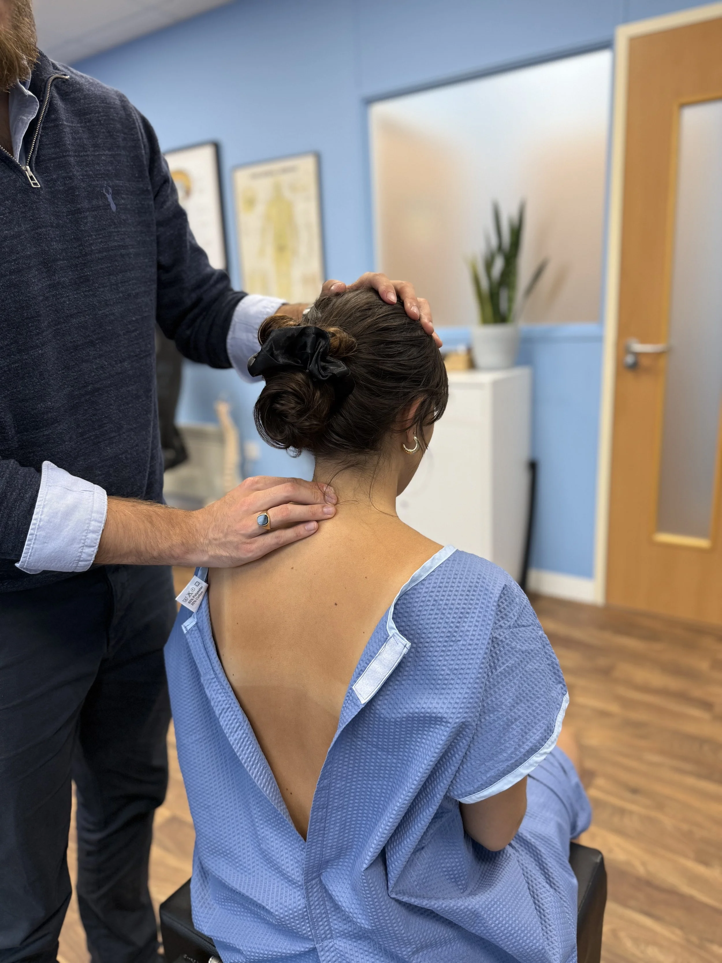 A woman in a blue gown receiving a neck palpation from a Chiropractor in a dark sweater in a room with blue walls and framed posters.