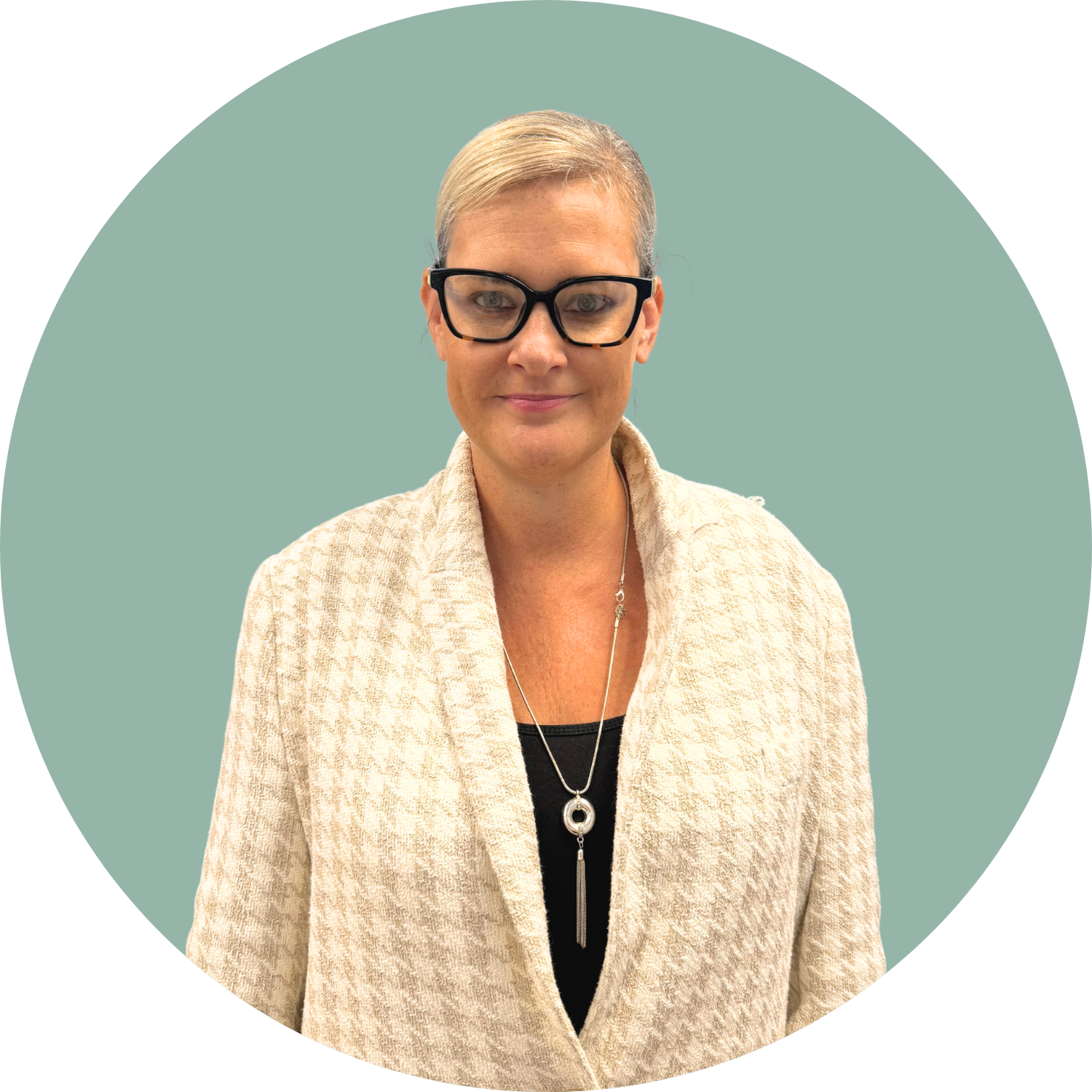 A woman with blonde hair wearing glasses, a beige textured blazer, a black top, and jewelry, standing against a muted green background.